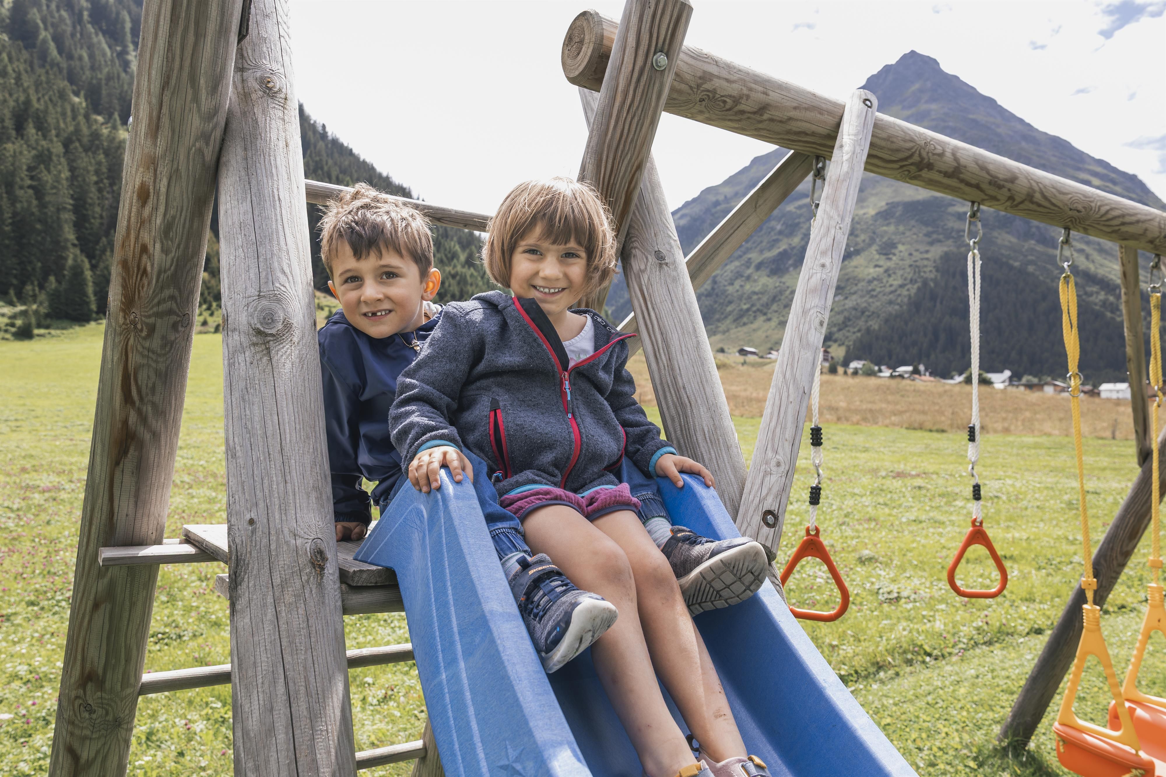Zwei Kinder sitzen fröhlich auf einer Rutsche in einem Spielplatz. Im Hintergrund sind grüne Wiesen und Berge zu sehen.