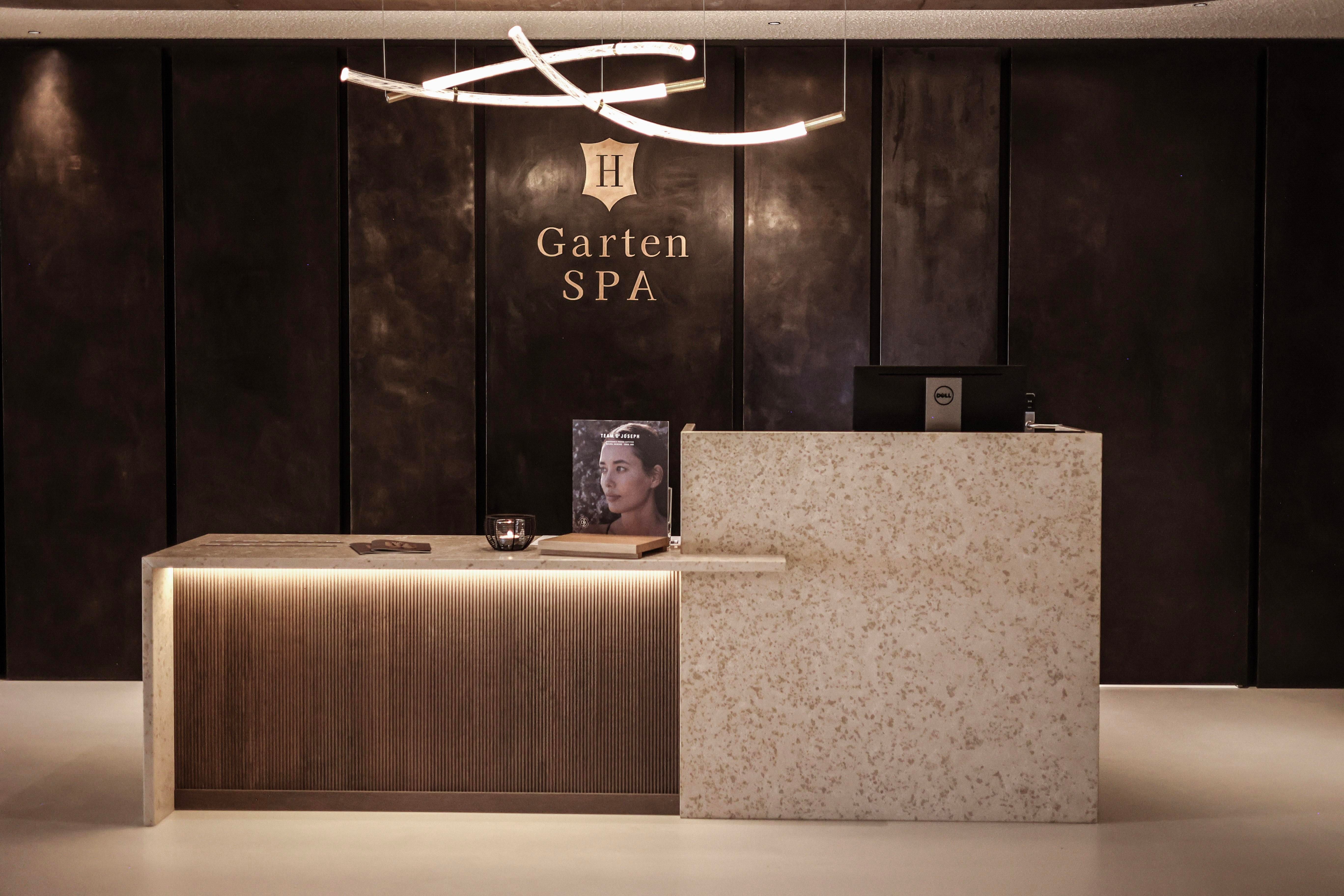 Ein modernes Empfangsbereich des Gartens SPA mit einem eleganten Tresen aus Holz und Stein. Die Wände sind dunkel und stilvoll, und eine sanfte Beleuchtung sorgt für eine entspannte Atmosphäre.