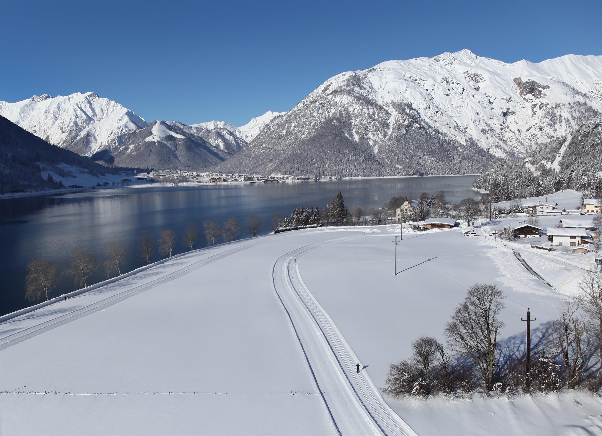 Eine verschneite Landschaft mit Bergen im Hintergrund und einem klaren blauen Himmel. Ein See spiegelt die Umgebung wider und sorgt für eine ruhige Atmosphäre.