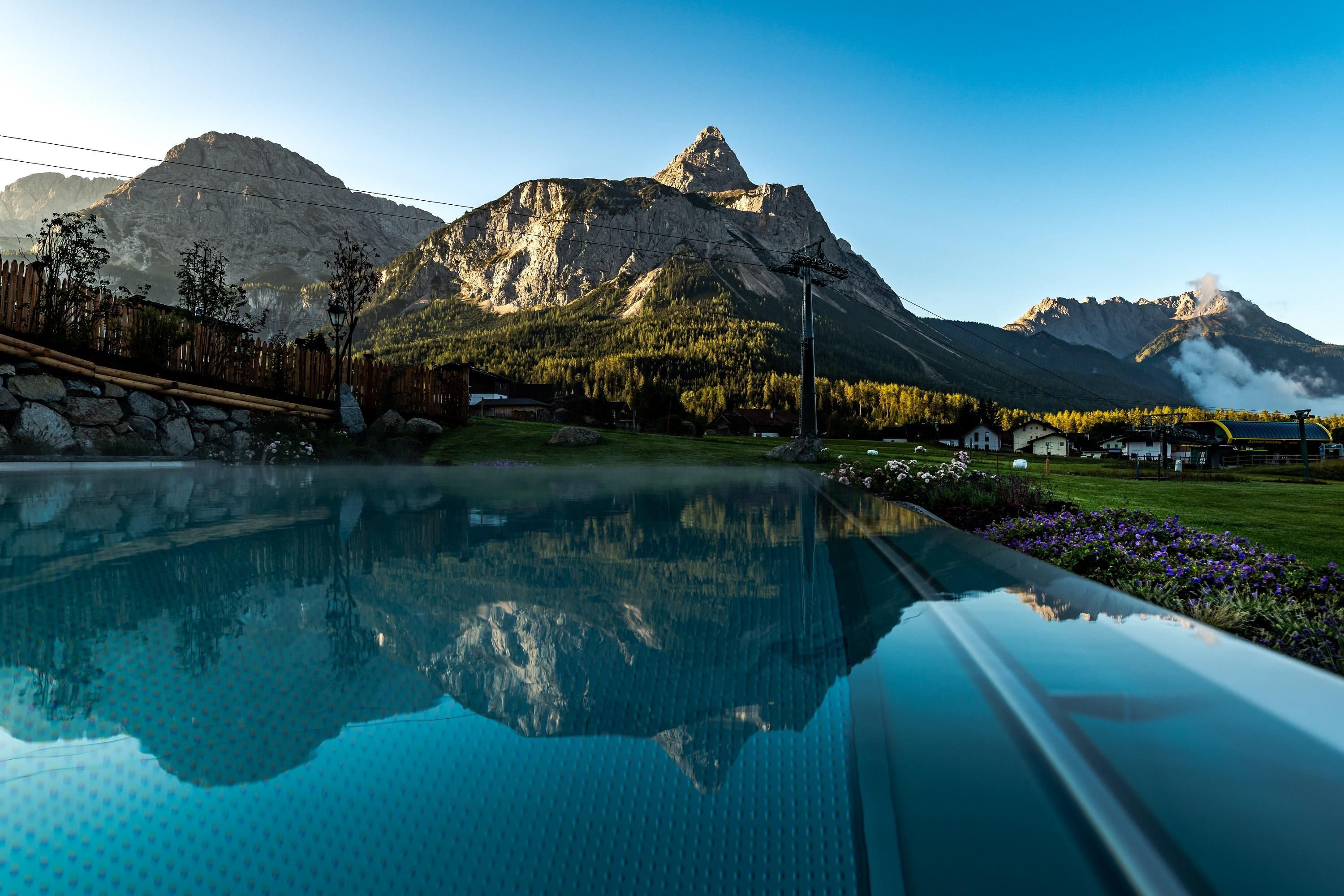 Ein ruhiger Pool mit klarer Wasseroberfläche spiegelt die majestätischen Berge im Hintergrund wider. Die Landschaft ist von sanften Hügeln und Bäumen umgeben.