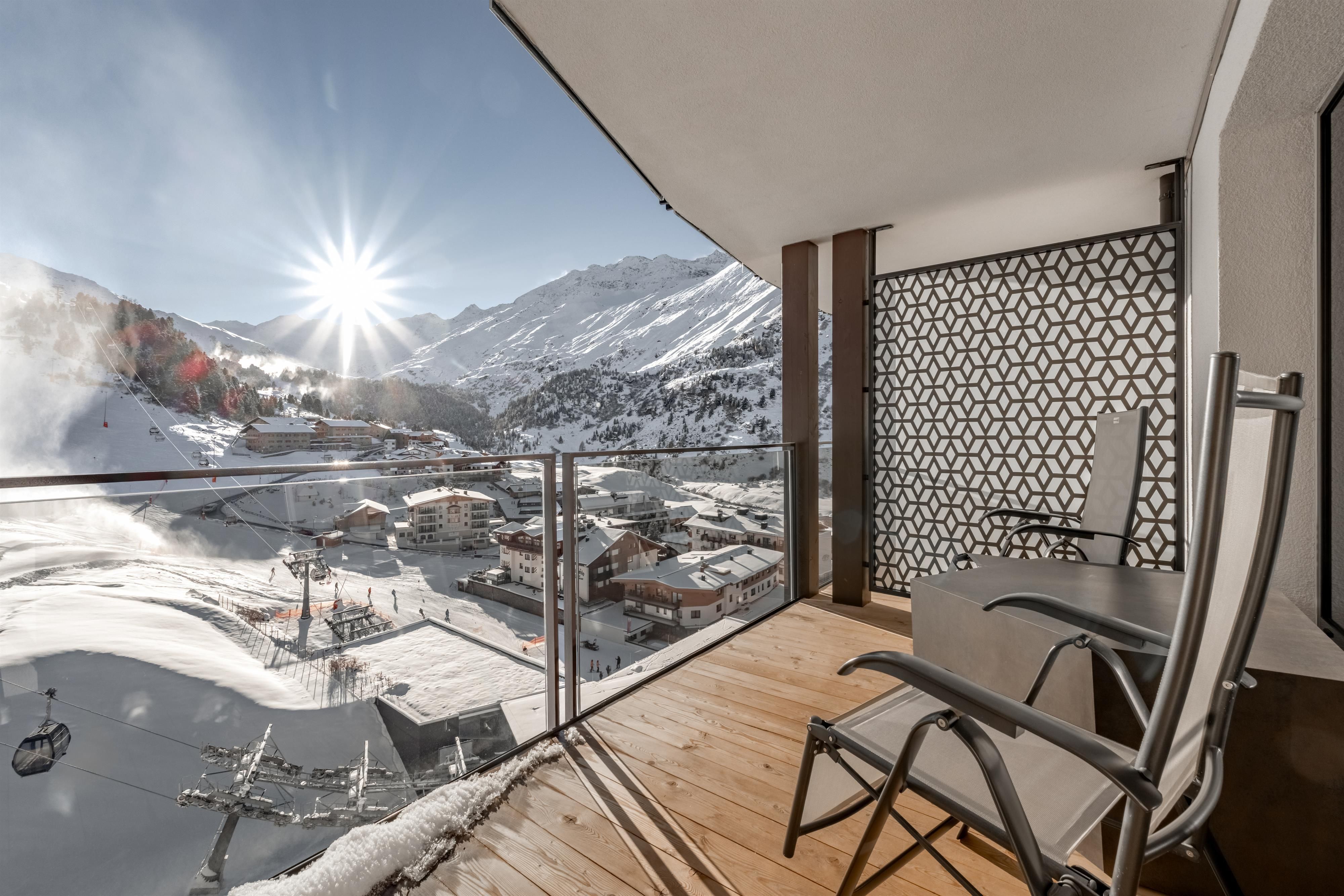 Ein Balkon mit Holzfußboden und zwei Stühlen. Im Hintergrund erstrecken sich schneebedeckte Berge und die Sonne scheint hell.