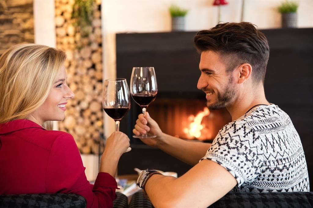Ein lächelndes Paar stößt mit Rotwein zusammen. Im Hintergrund brennt ein gemütliches Feuer.
