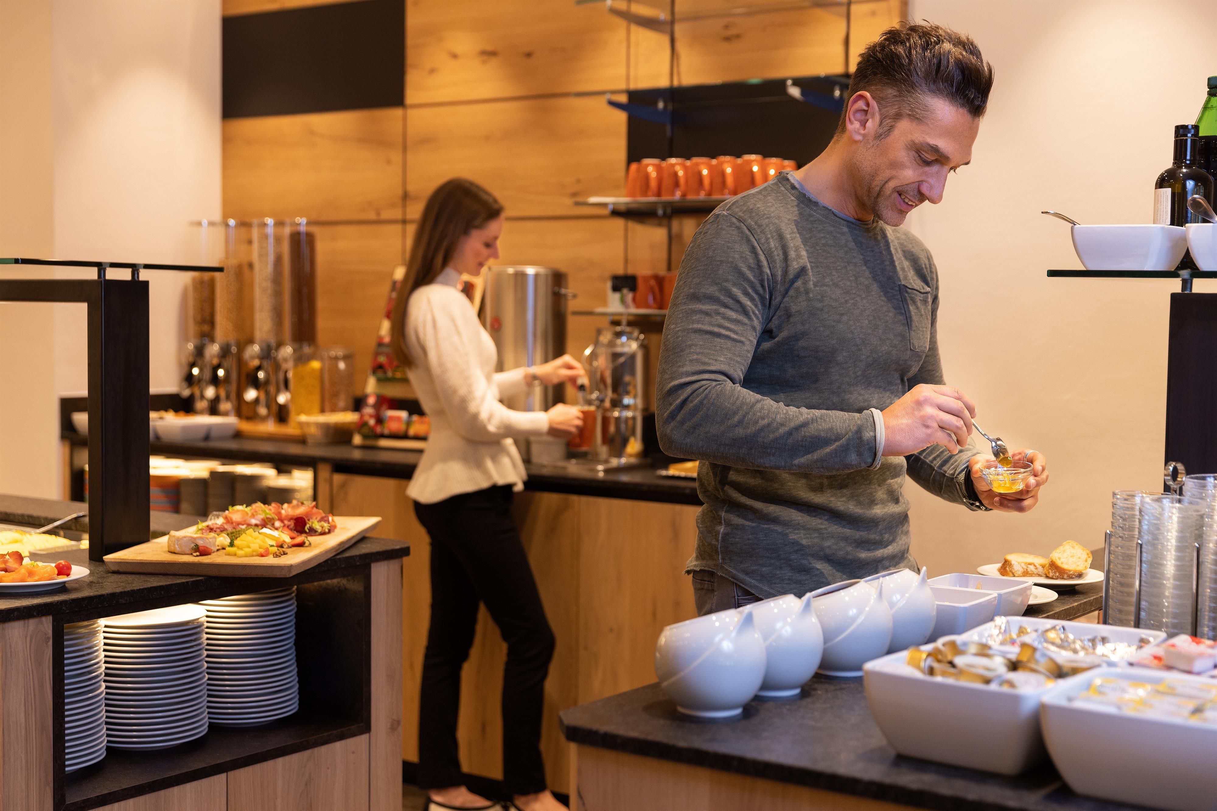 Eine moderne Frühstücksbar mit verschiedenen Speisen und Getränken. Zwei Personen bereiten sich auf das Frühstück vor.