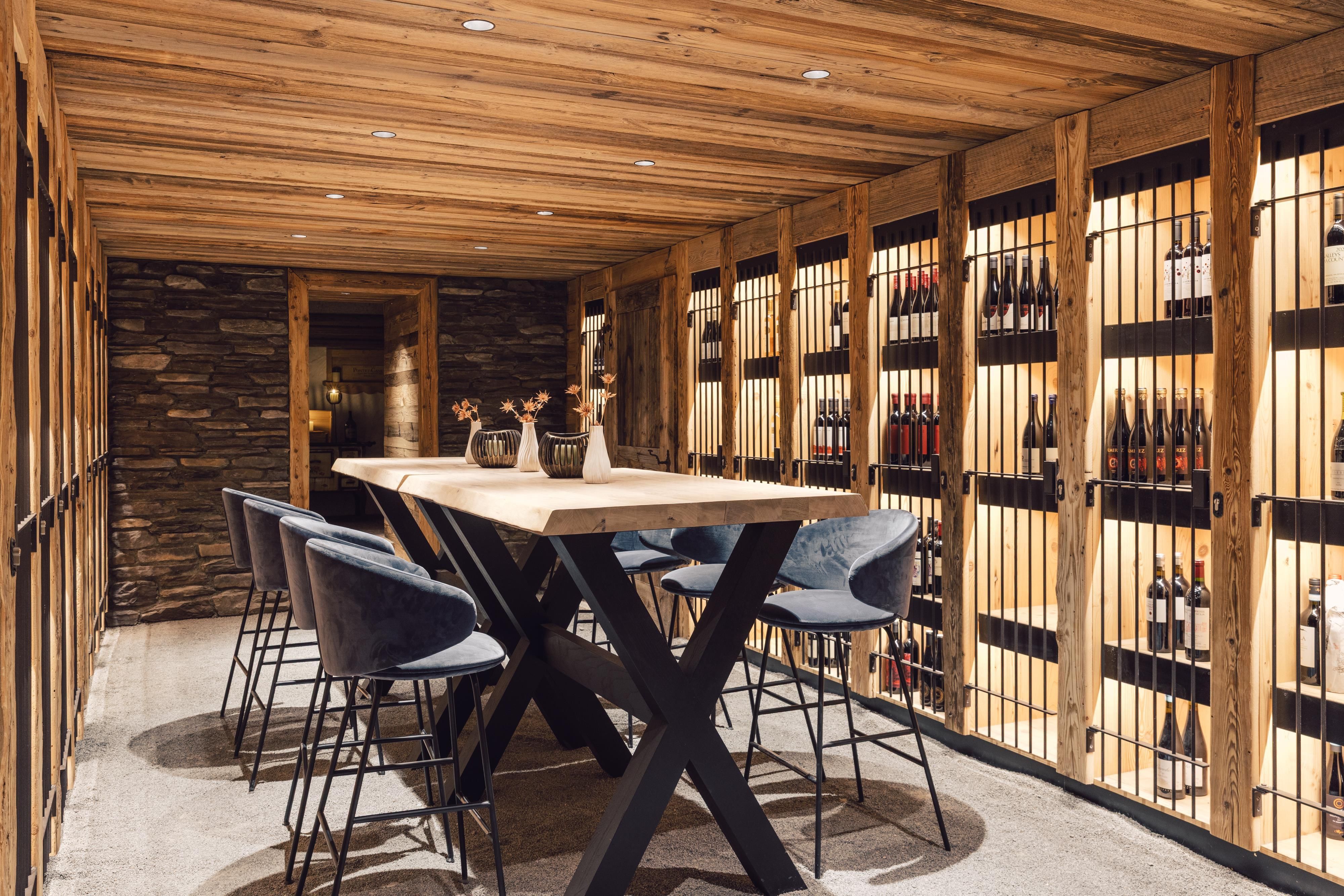 Ein stilvoller Weinkeller mit Holzdecke und gestreiften Regalen. Ein großer Tisch mit Stühlen lädt zum Verweilen ein.