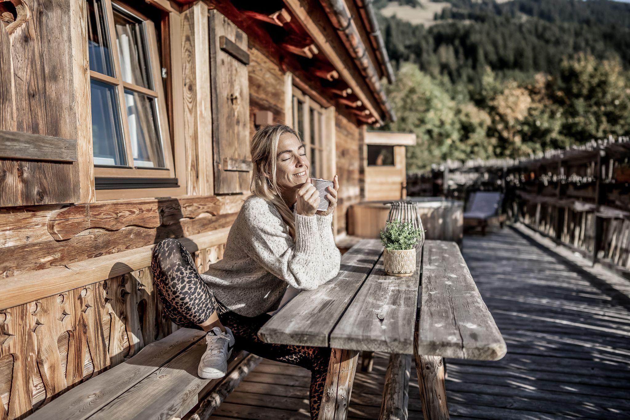 Eine Frau sitzt entspannt an einem Holztisch vor einer gemütlichen Chalet. Sie genießt eine Tasse Kaffee im Freien, umgeben von einer schönen Landschaft.
