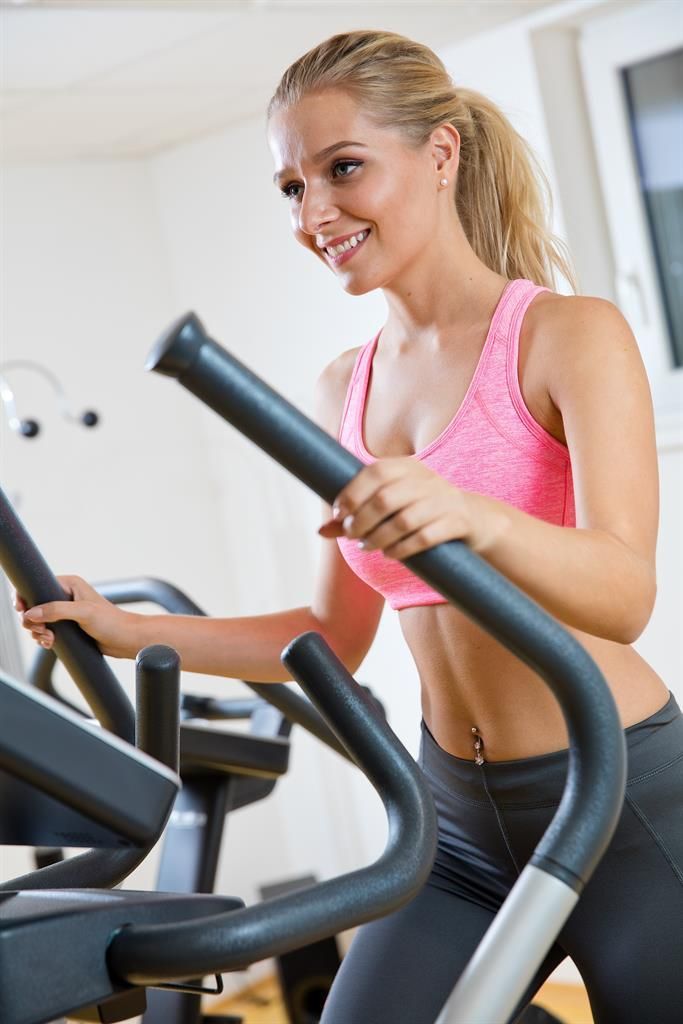 Eine fröhliche Frau trainiert auf einem Ellipsentrainer in einem modernen Fitnessstudio. Sie trägt ein pinkes Sporttop und lächelt.