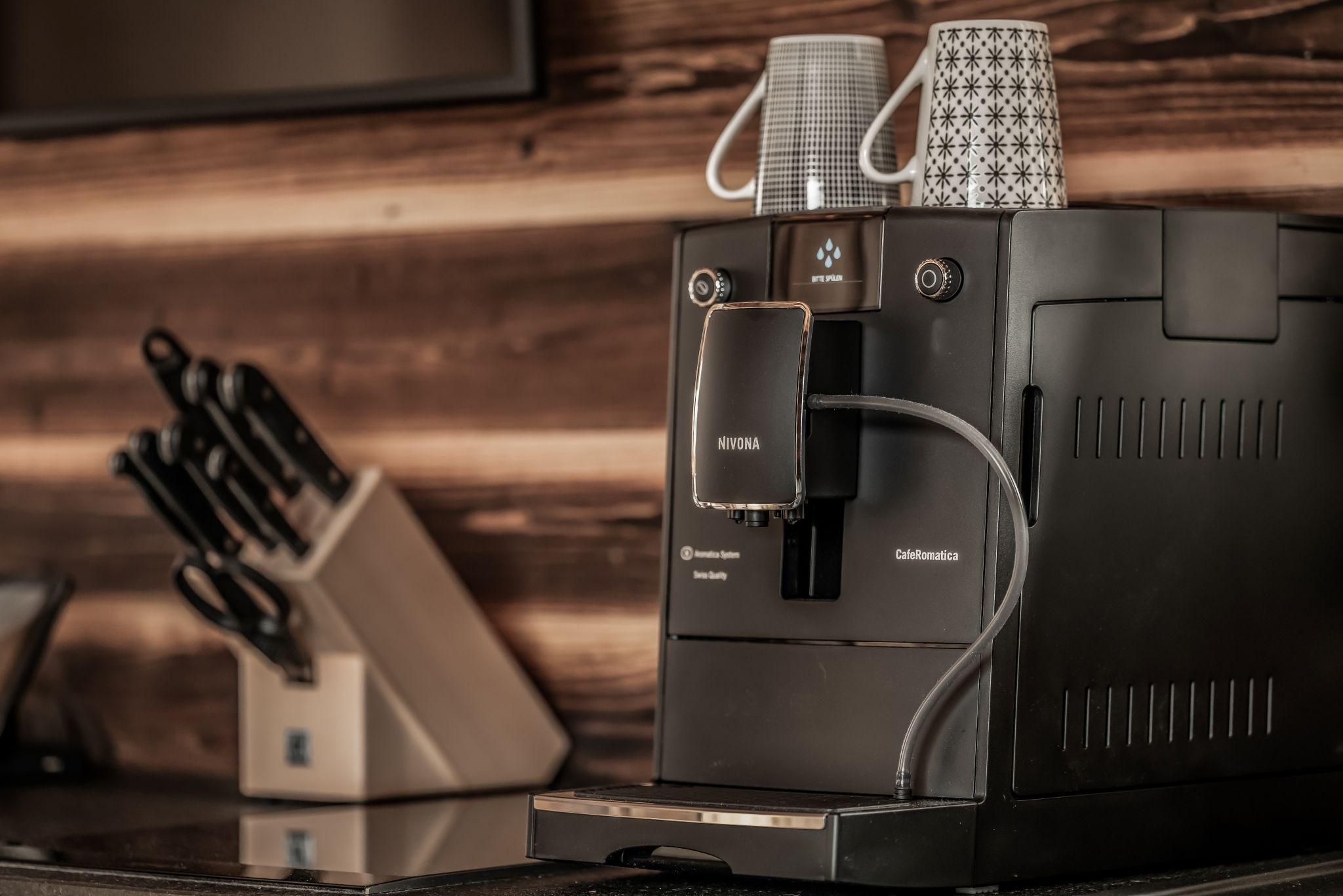 Eine moderne Kaffeemaschine steht auf einer Arbeitsfläche. Im Hintergrund sind zwei Tassen und ein Messerblock zu sehen.