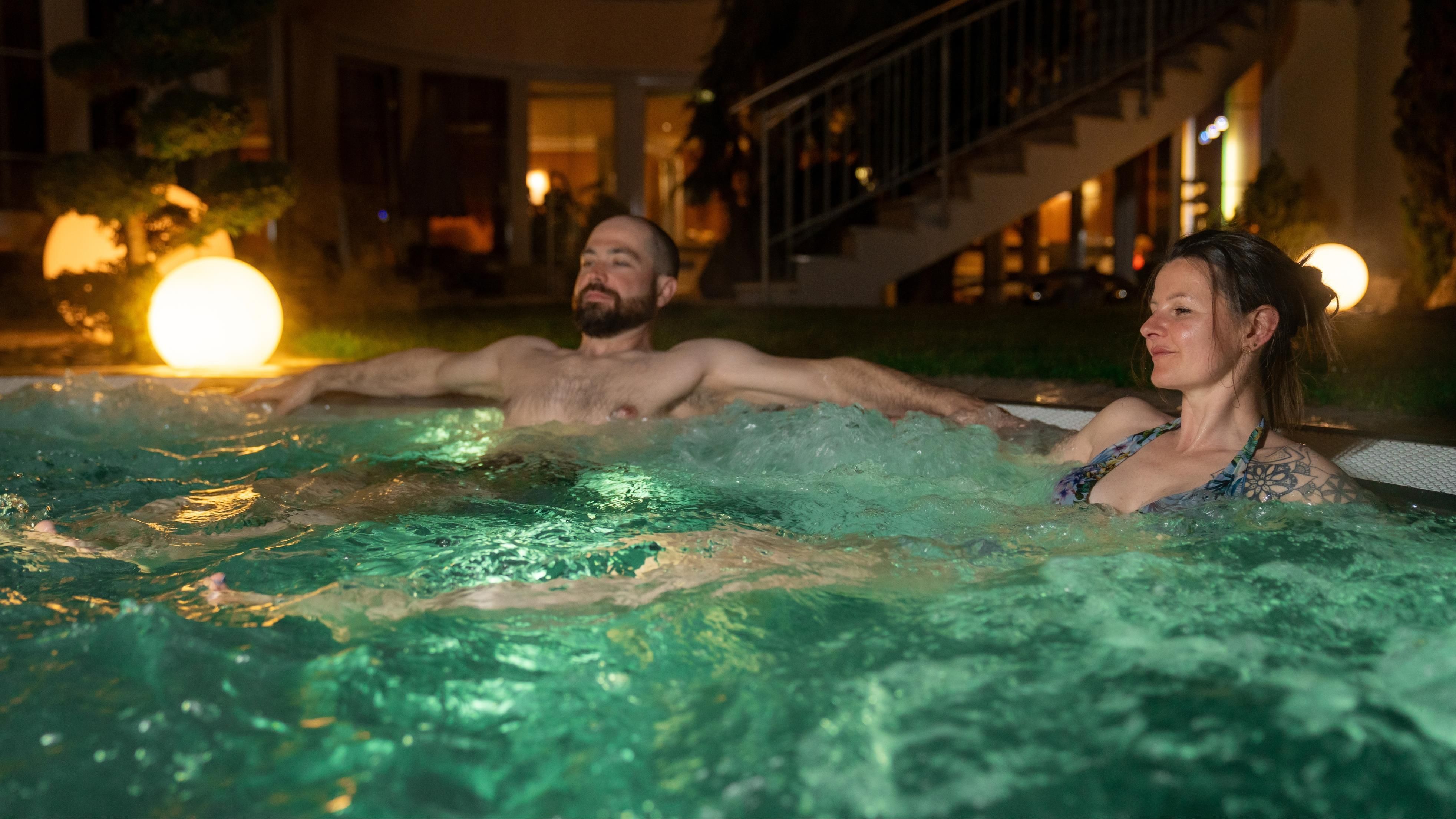Ein Paar entspannt im warmen Wasser eines Whirlpools. Der Abend ist gemütlich mit sanften Lichtern und einer ruhigen Atmosphäre.