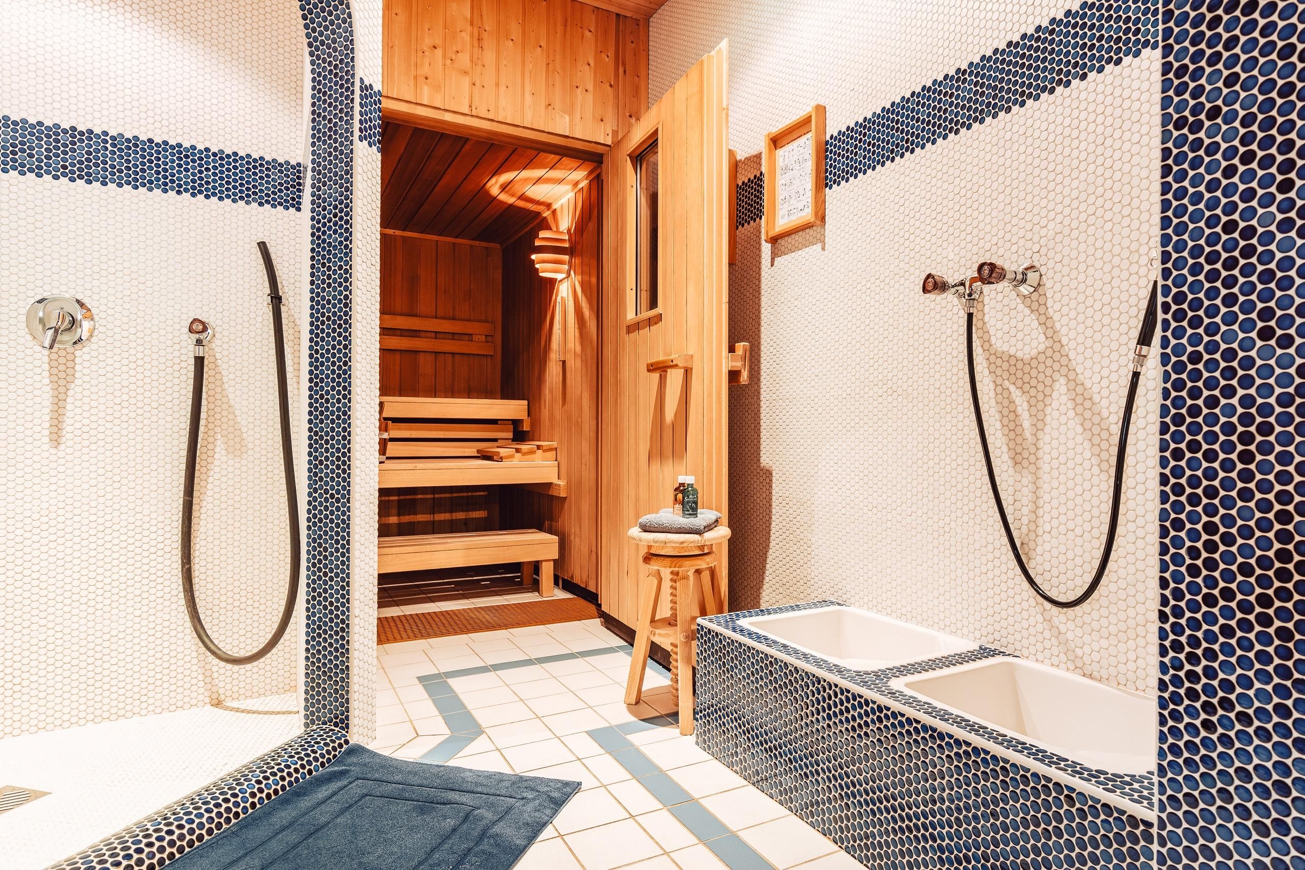 Ein modernes Badezimmer mit einer Sauna und schönen blauen Fliesen. Zwei Duschbereiche und ein Holzstuhl runden das entspannende Ambiente ab.