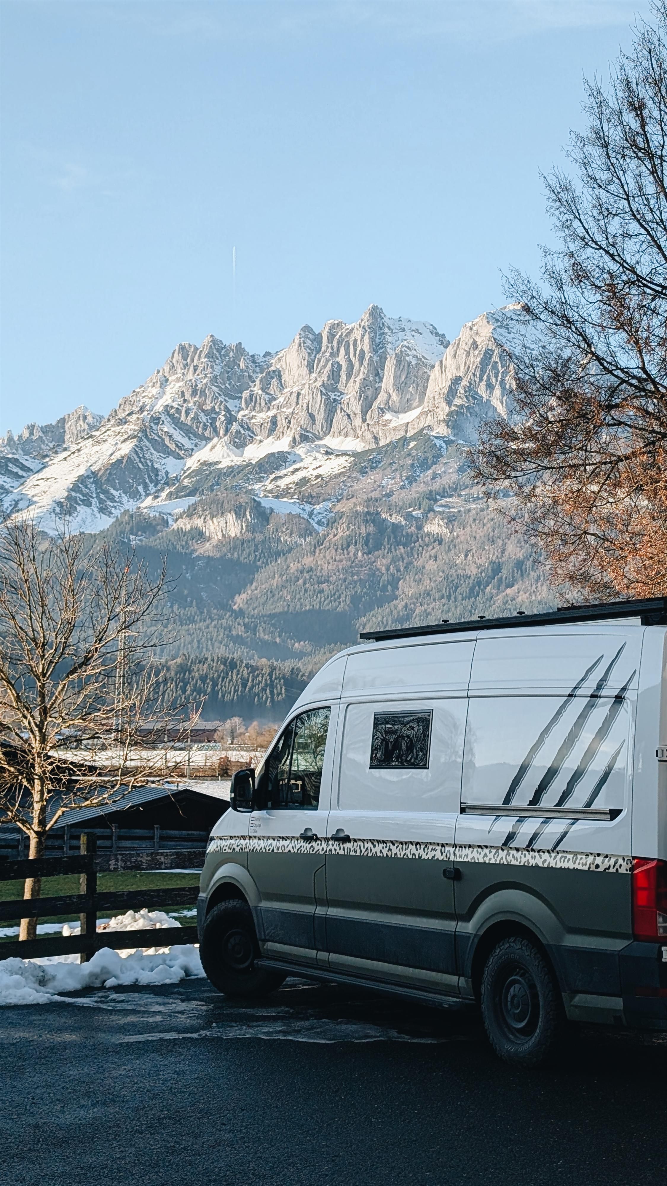 Ein Wohnmobil steht auf einem Parkplatz mit Blick auf majestätische Berge. Die Landschaft ist schneebedeckt und der Himmel ist klar.