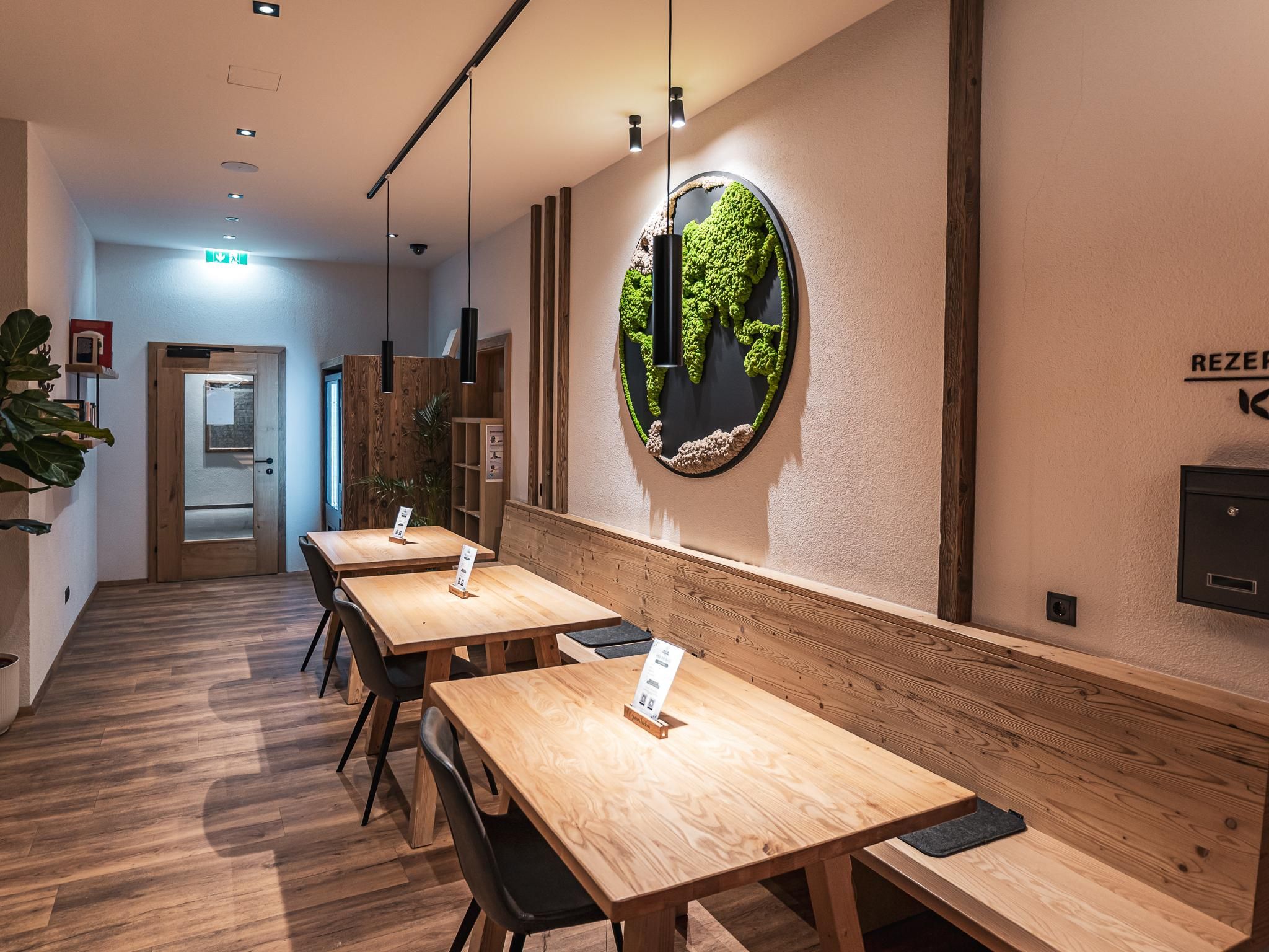 Ein modern eingerichteter Raum mit Holztischen und Stühlen. Eine grüne Wanddekoration und sanfte Beleuchtung schaffen eine einladende Atmosphäre.