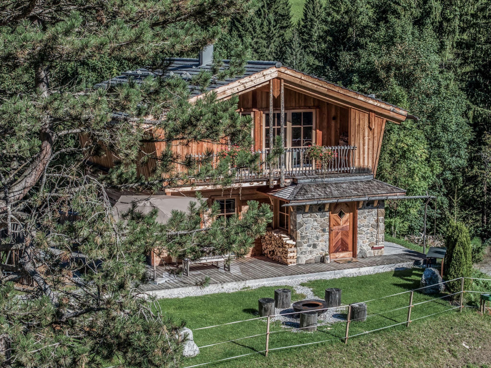 Ein schönes Chalet umgeben von Bäumen und gepflegtem Grün. Vor dem Haus befindet sich eine Feuerstelle mit Sitzmöglichkeiten.