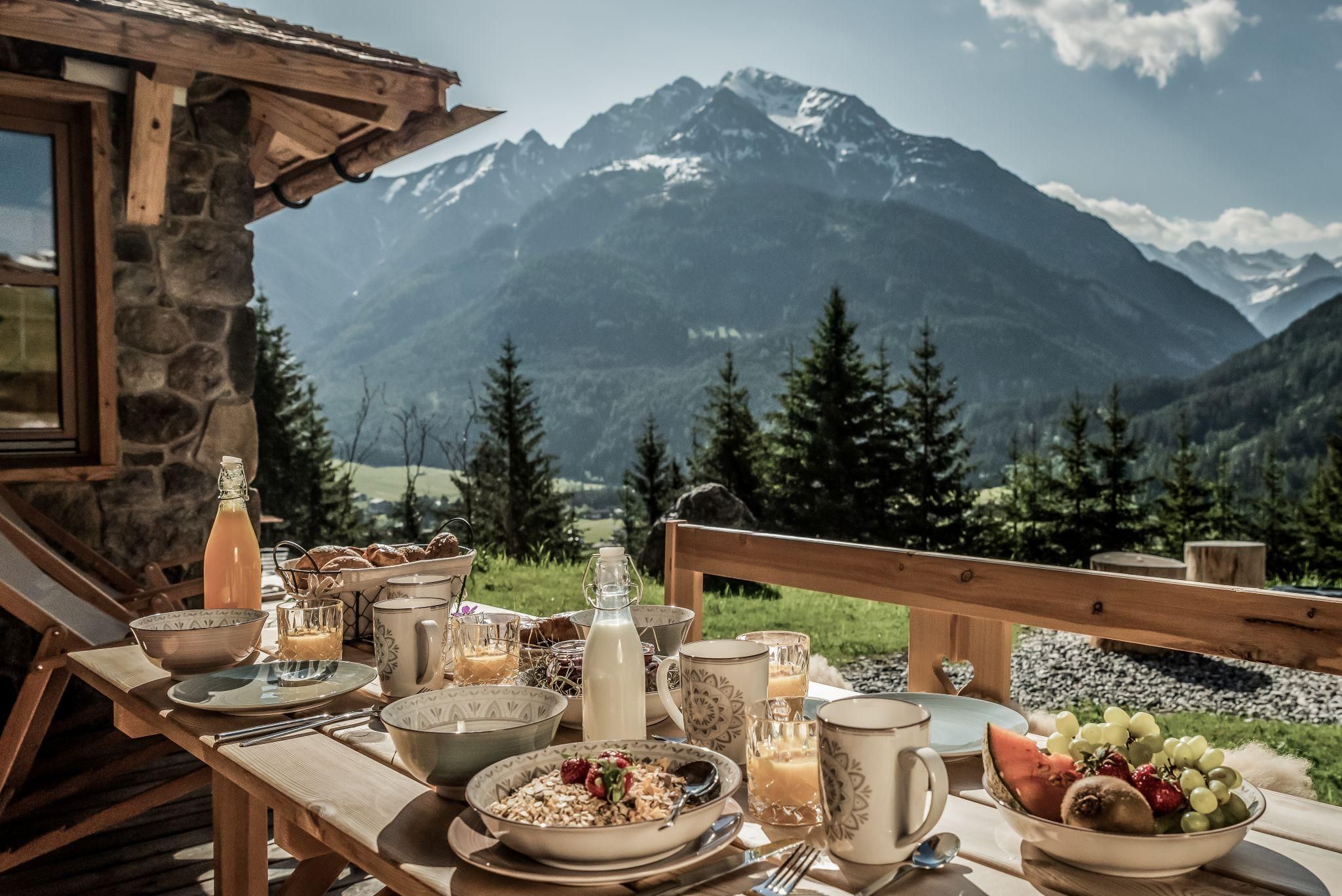 Ein gedeckter Tisch mit einem reichhaltigen Frühstück steht auf einer Terrasse. Im Hintergrund sind majestätische Berge und ein klarer Himmel zu sehen.