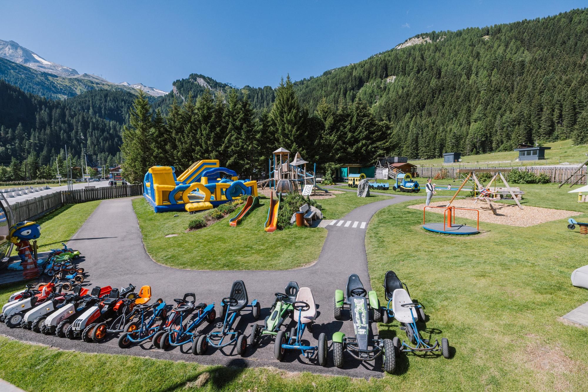 Ein Spielplatz mit verschiedenen Attraktionen und einem Bereich für Kinderfahrzeuge. Im Hintergrund sind grüne Berge und ein strahlend blauer Himmel zu sehen.