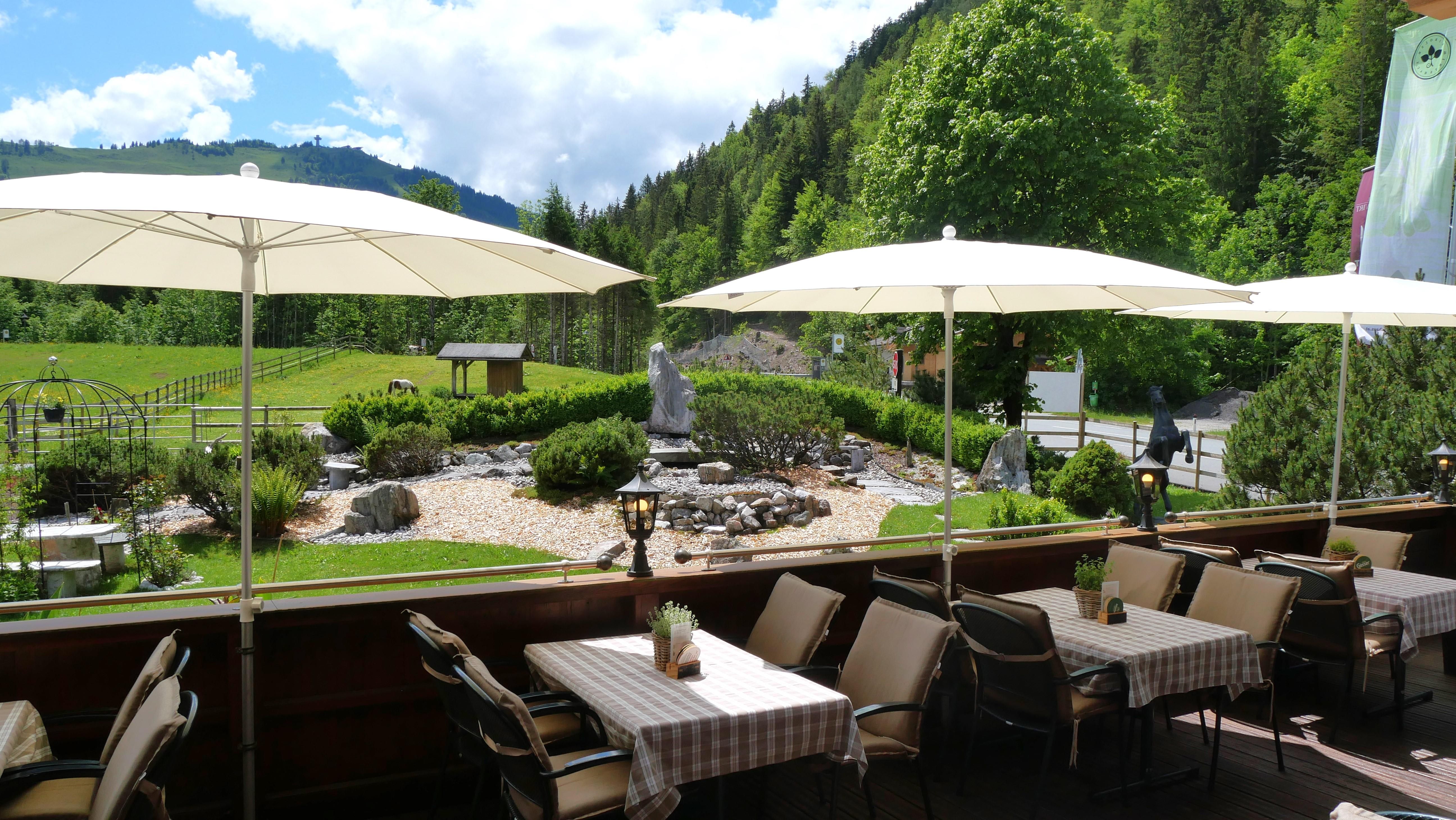Eine gemütliche Terrasse mit Tischdecken und Sonnenschirmen. Im Hintergrund sind grüne Bäume und eine gepflegte Gartenlandschaft zu sehen.