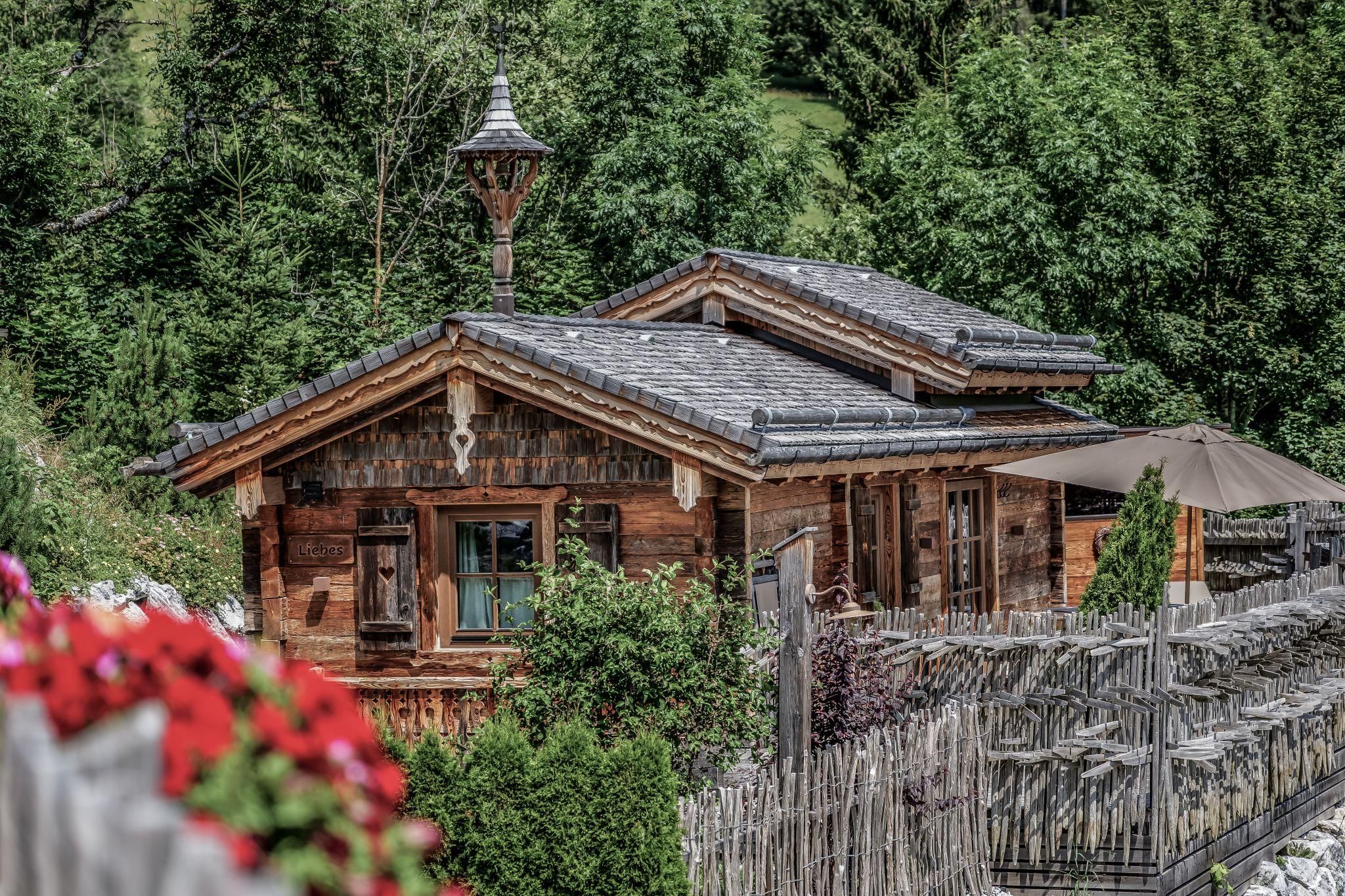 Ein idyllisches Chalet umgeben von grünen Bäumen. Blumen und ein Sonnenschirm verleihen der Szene einen einladenden Charme.