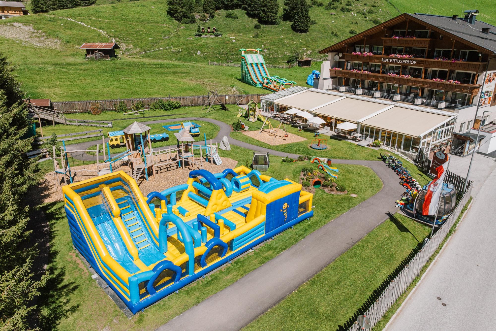 Ein großer, bunter Hüpfburg-Spielplatz mit Rutschen und verschiedenen Spielgeräten. Im Hintergrund ist ein Hotel mit Terrasse und Grünflächen zu sehen.