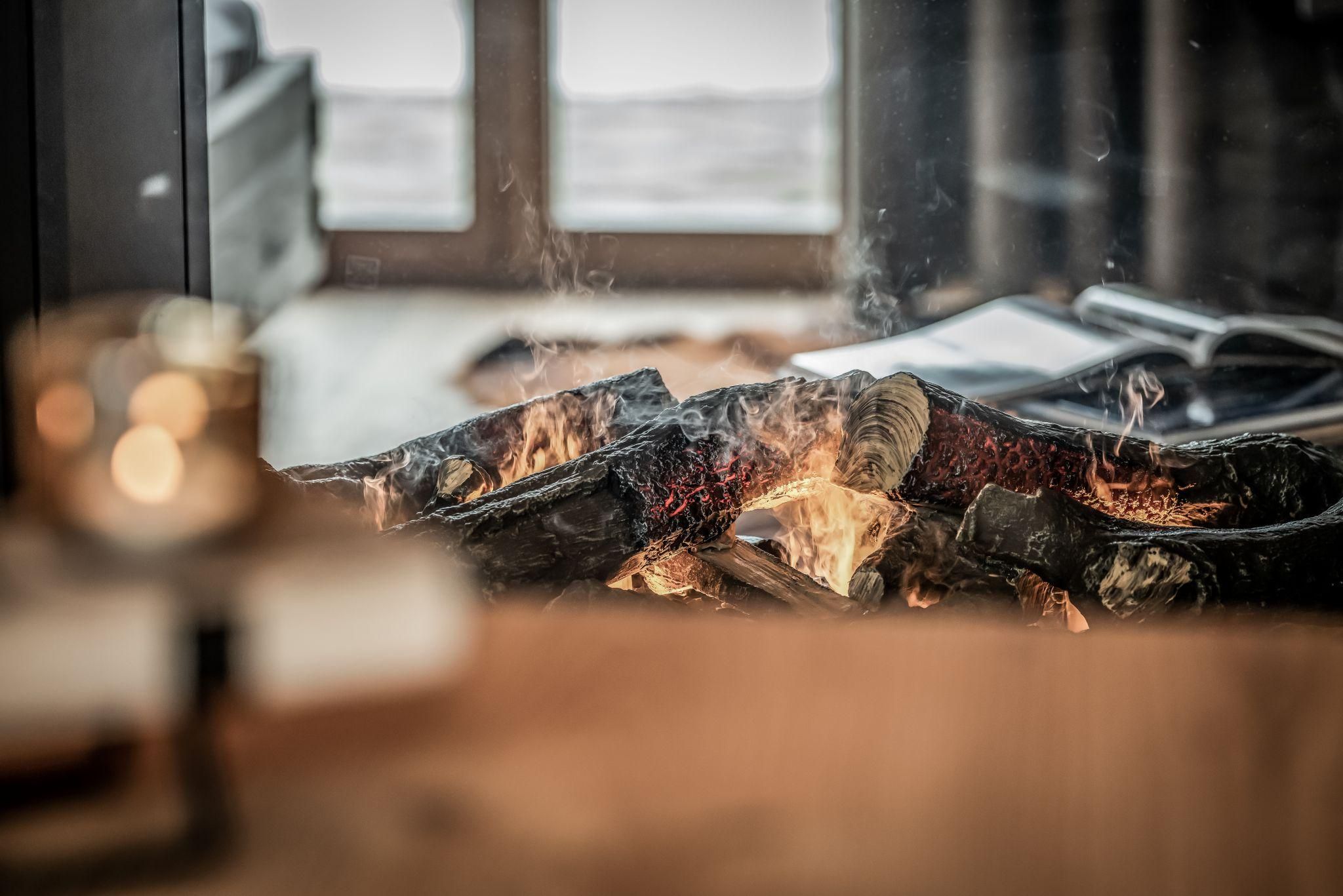 Ein gemütlicher Kamin mit brennendem Feuer und glühenden Holzscheiten. Im Hintergrund sind Fenster zu sehen, die Tageslicht hereinlassen.