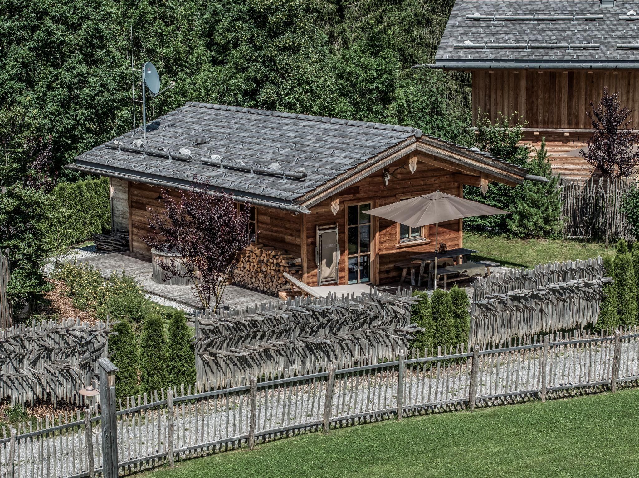 Ein gemütliches Chalet mit einem Sonnenschirm und einer Sitzecke im Freien. Es ist von einem gepflegten Garten und einem Zaun umgeben.
