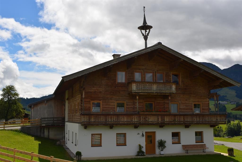 Ein traditionelles Holzhaus in den Alpen mit einem schönen Balkon. Die Umgebung ist grün und die Berge sind im Hintergrund sichtbar.