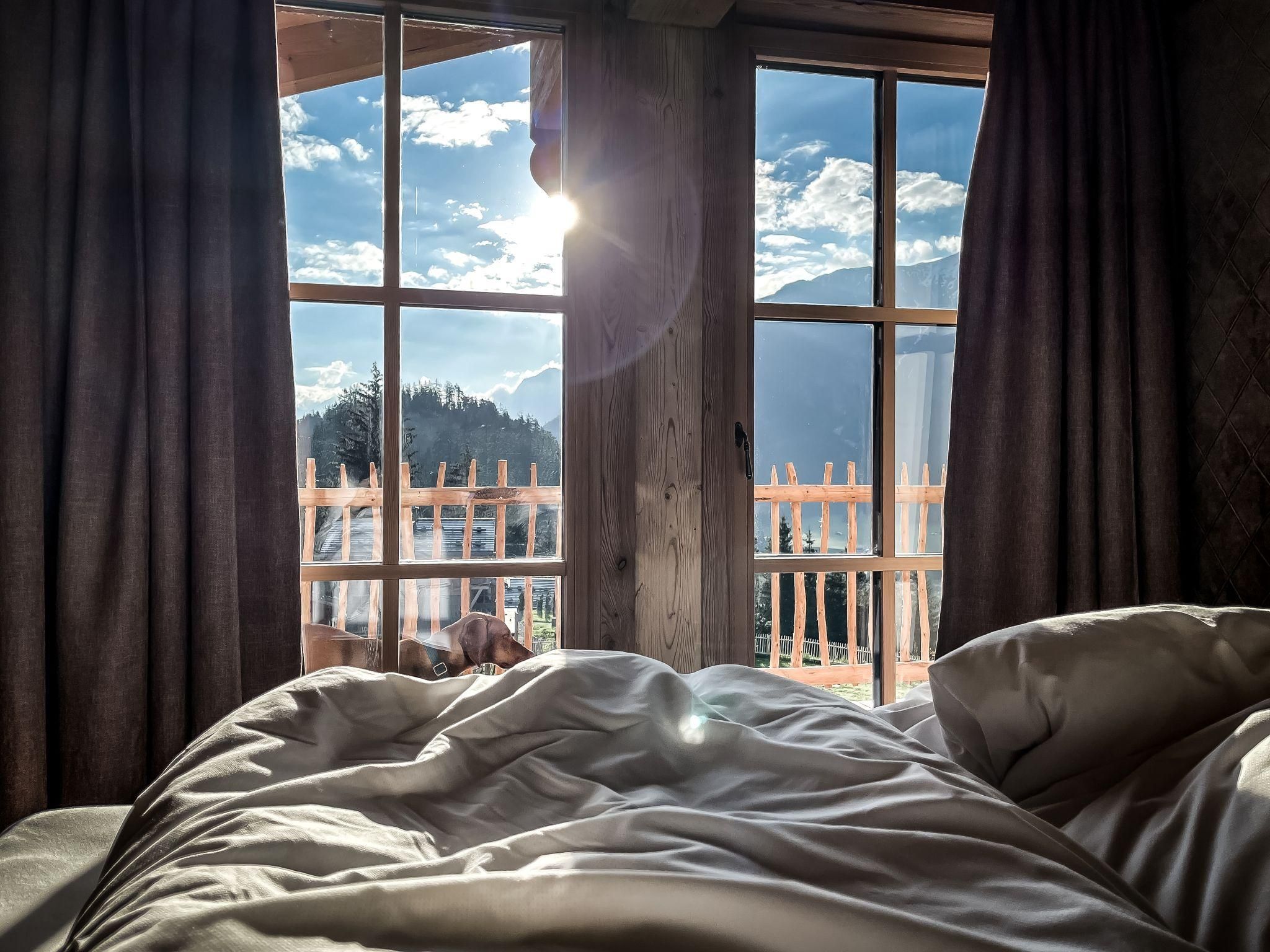 Ein gemütliches Bett mit weißen Bettlaken und einem strahlenden Sonnenlicht, das durch das Fenster scheint. Draußen sind Berge und ein blauer Himmel sichtbar.