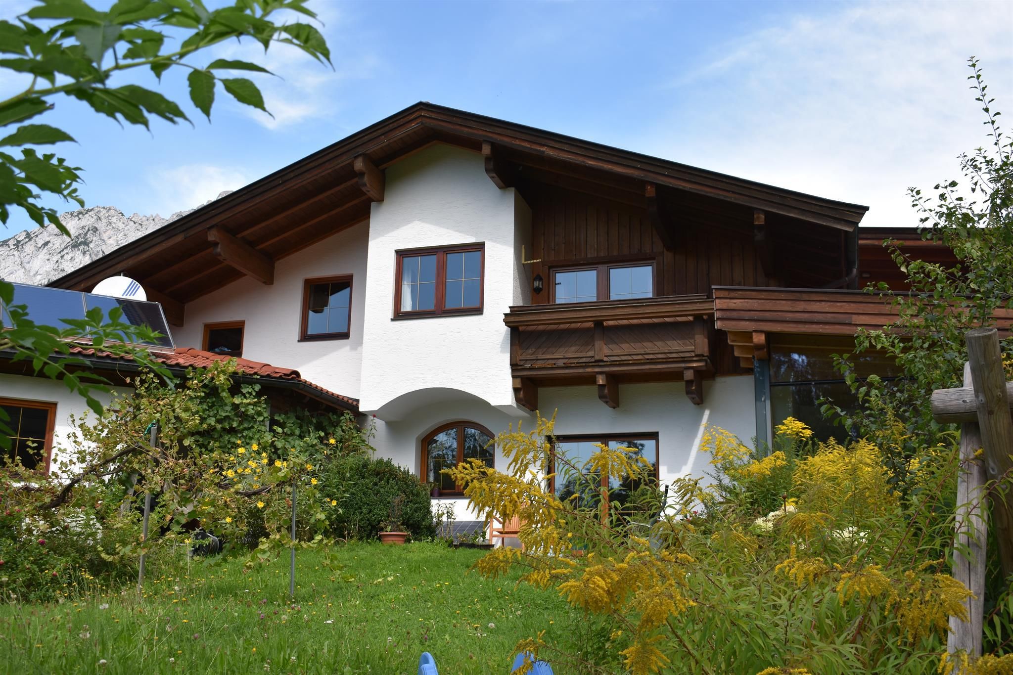 Ein charmantes Haus mit Holzverkleidung und großen Fenstern in einem grünen Garten. Im Hintergrund sind sanfte Hügel sichtbar.