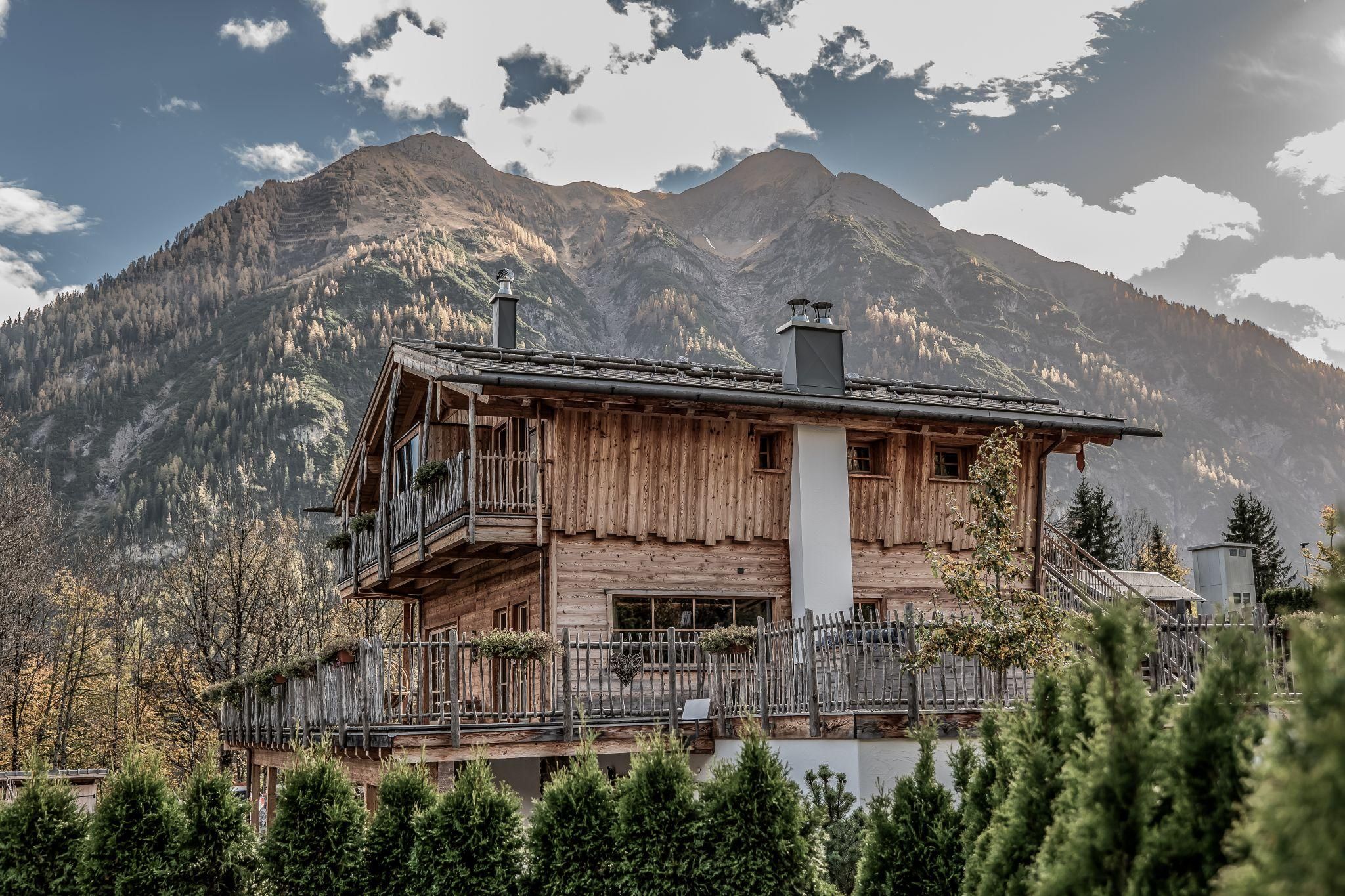 Ein malerisches Chalet mit Balkon, umgeben von grünen Bäumen. Im Hintergrund erhebt sich ein majestätisches Gebirge unter einem teilweise bewölkten Himmel.