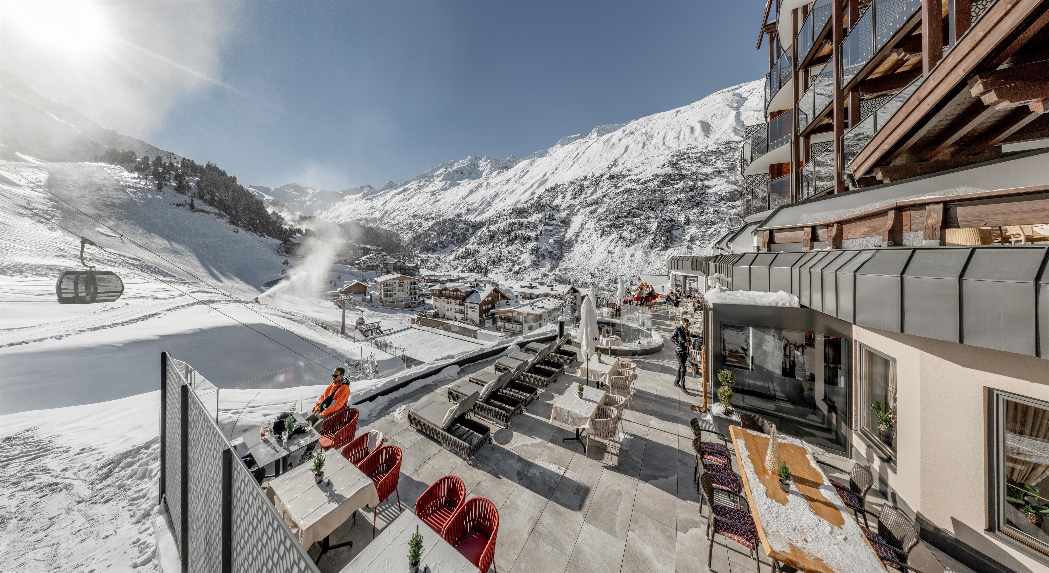 Ein wunderschöner Blick auf eine alpine Landschaft mit schneebedeckten Bergen. Im Vordergrund sieht man gemütliche Sitzgelegenheiten und eine Seilbahn, die die Gebirgswelt erschließt.