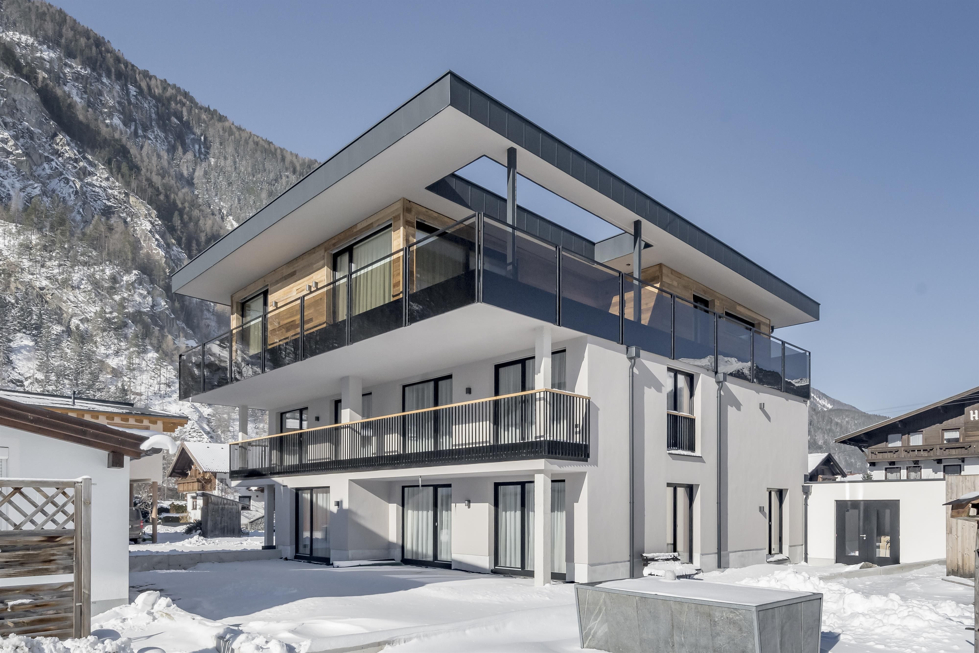 Ein modernes Gebäude mit mehreren Balkonen in einem schneebedeckten Landschaftszug. Im Hintergrund sind Berge zu sehen und der Himmel ist klar.