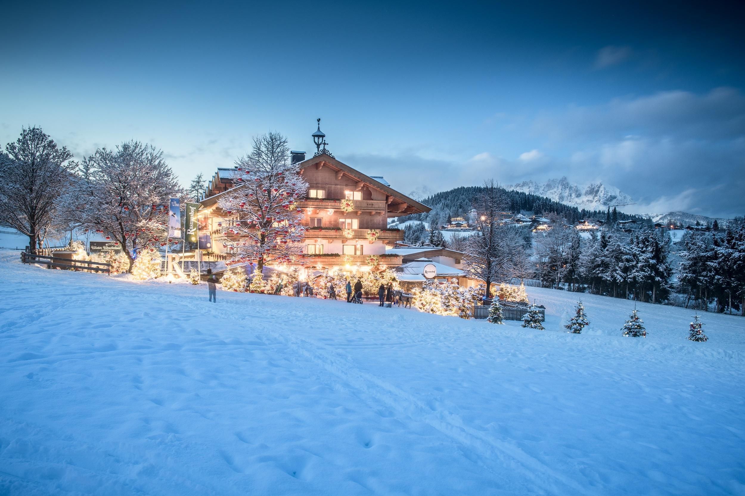Ein wunderschönes, winterliches Haus mit Weihnachtslichtern in einer verschneiten Landschaft. Bäume und schneebedeckte Hügel umgeben die Szenerie.