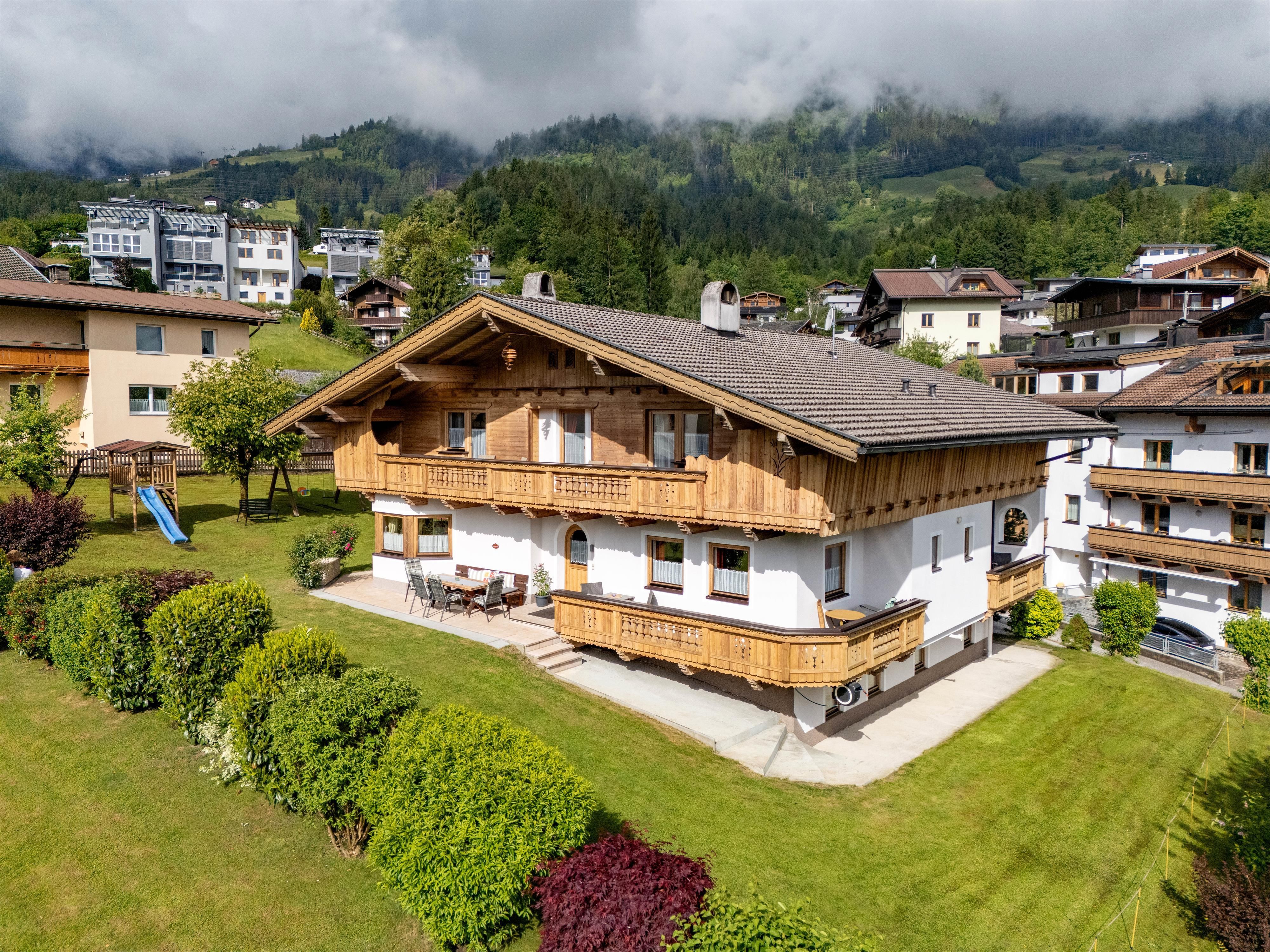 Ein charmantes Alpenhaus mit Holzverkleidung in einer grünen Umgebung. Im Hintergrund sind Berge und andere Gebäude sichtbar.
