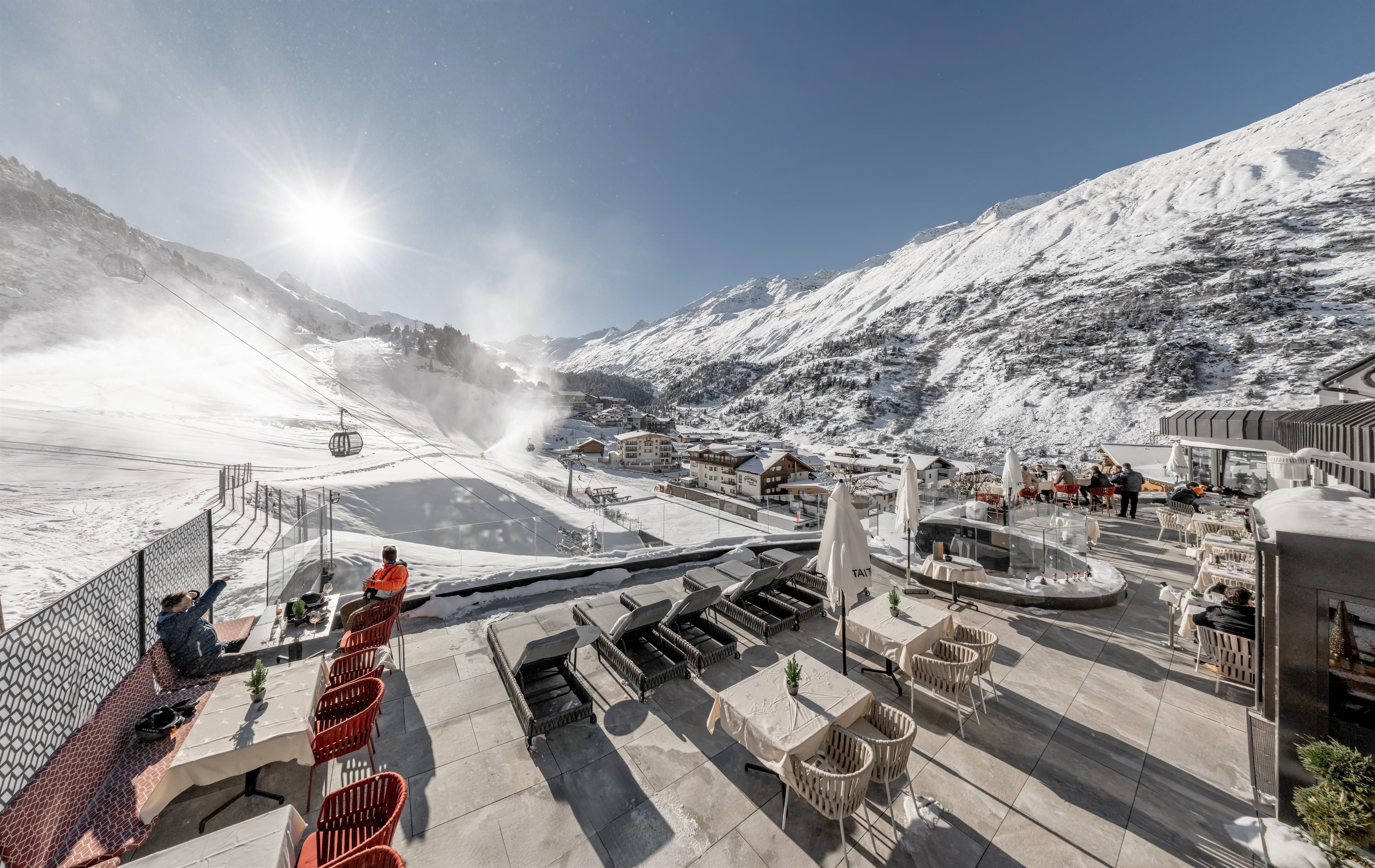 Eine Berglandschaft voller Schnee mit einem klaren blauen Himmel. Im Vordergrund sind Liegen und Sitzgelegenheiten auf einer Terrasse zu sehen.