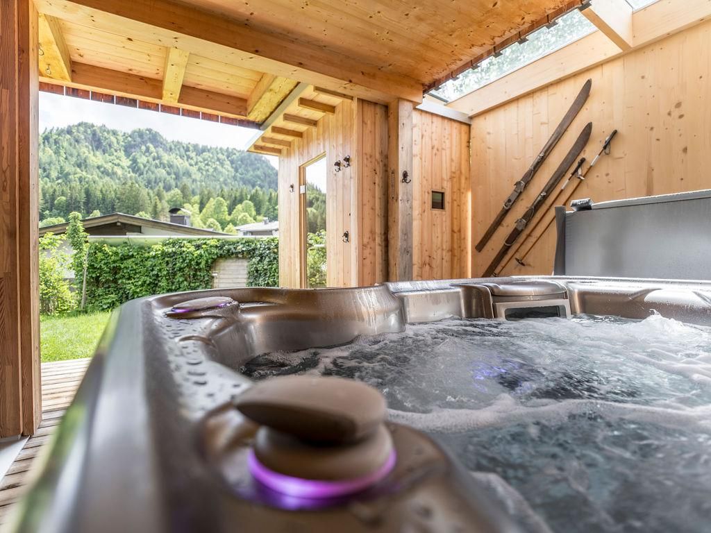 Ein gemütlicher Whirlpool in einem Holzraum mit Blick auf die Berge. Im Hintergrund sind Skistöcke und eine grüne Landschaft zu sehen.