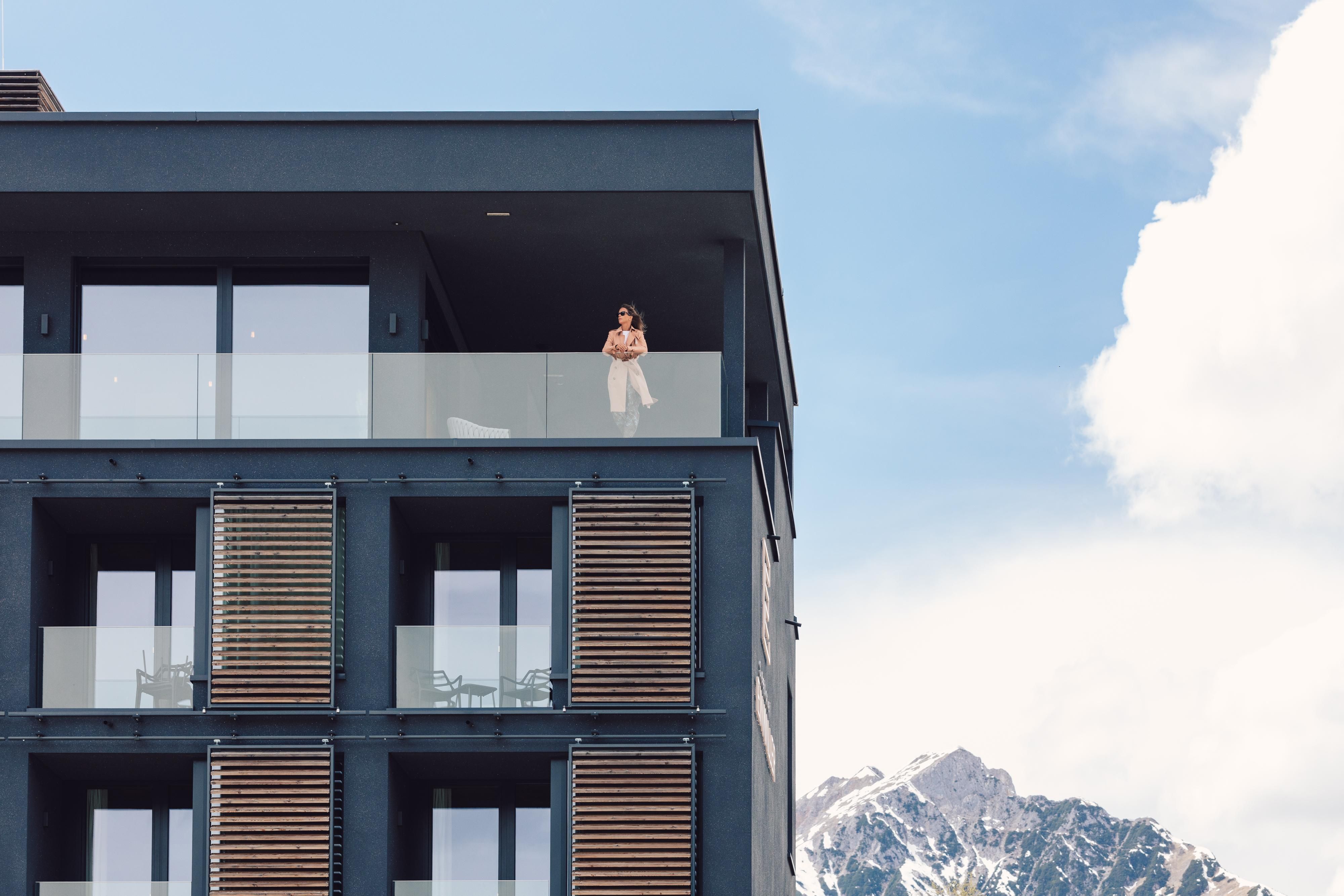 Ein modernes Gebäude mit Balkonen und großen Fenstern. Im Hintergrund sind beeindruckende Berge und ein blauer Himmel zu sehen.