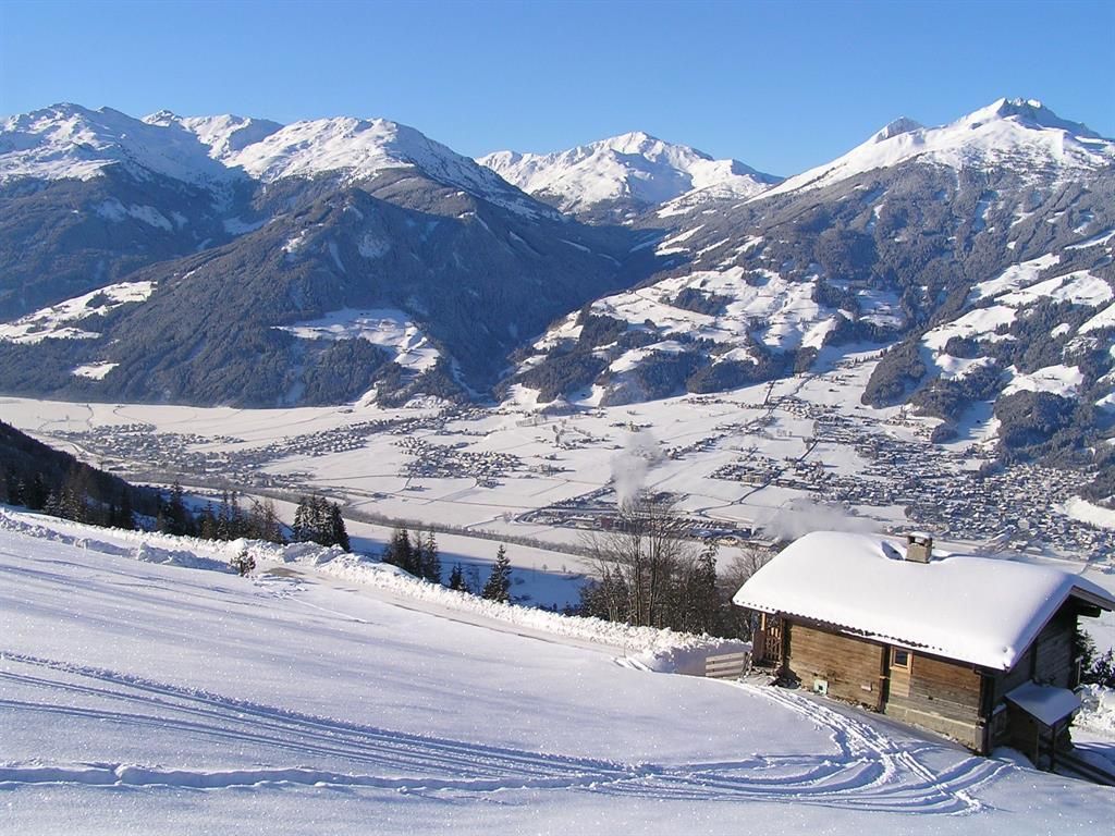 Eine wunderschöne Winterlandschaft mit schneebedeckten Bergen und einem kleinen Holzhaus im Vordergrund. Der Himmel ist klar und blau, während der Blick ins Tal, das mit Schnee bedeckt ist, fasziniert.