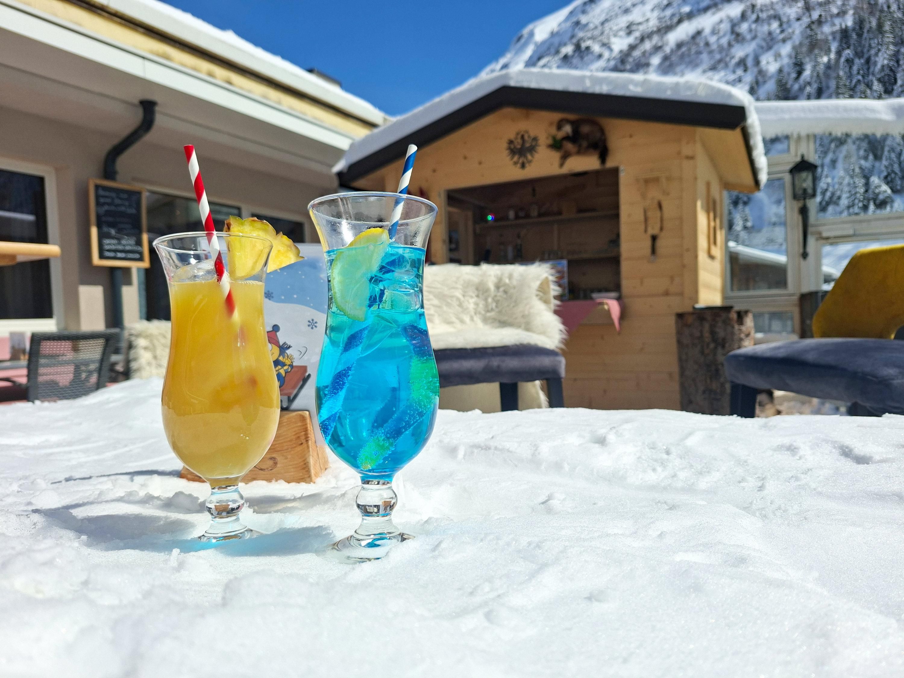 Zwei bunte Cocktails stehen auf dem Schnee. Im Hintergrund ist eine Chalet-Hütte und hohe Berge zu sehen.