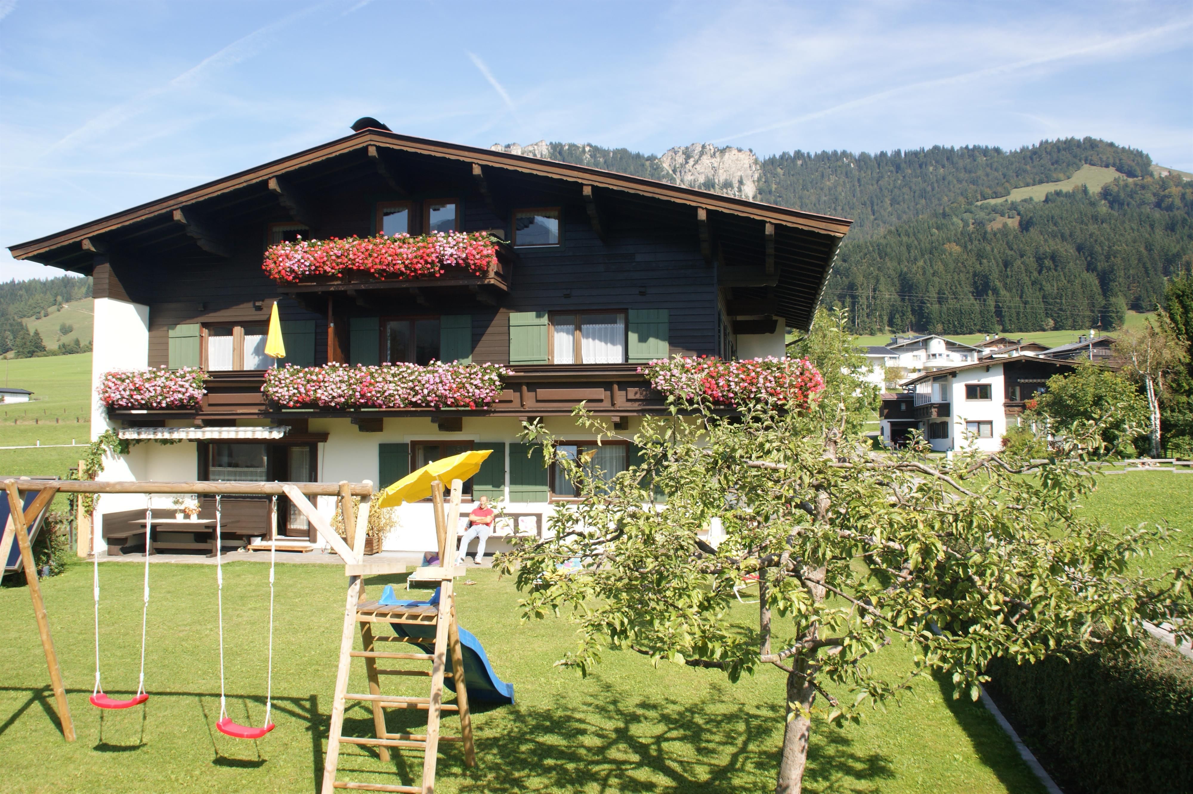 Ein schönes, mehrstöckiges Haus mit blühenden Fensterkästen. Im Vordergrund gibt es einen Garten mit einer Schaukel und einem Spielplatz.
