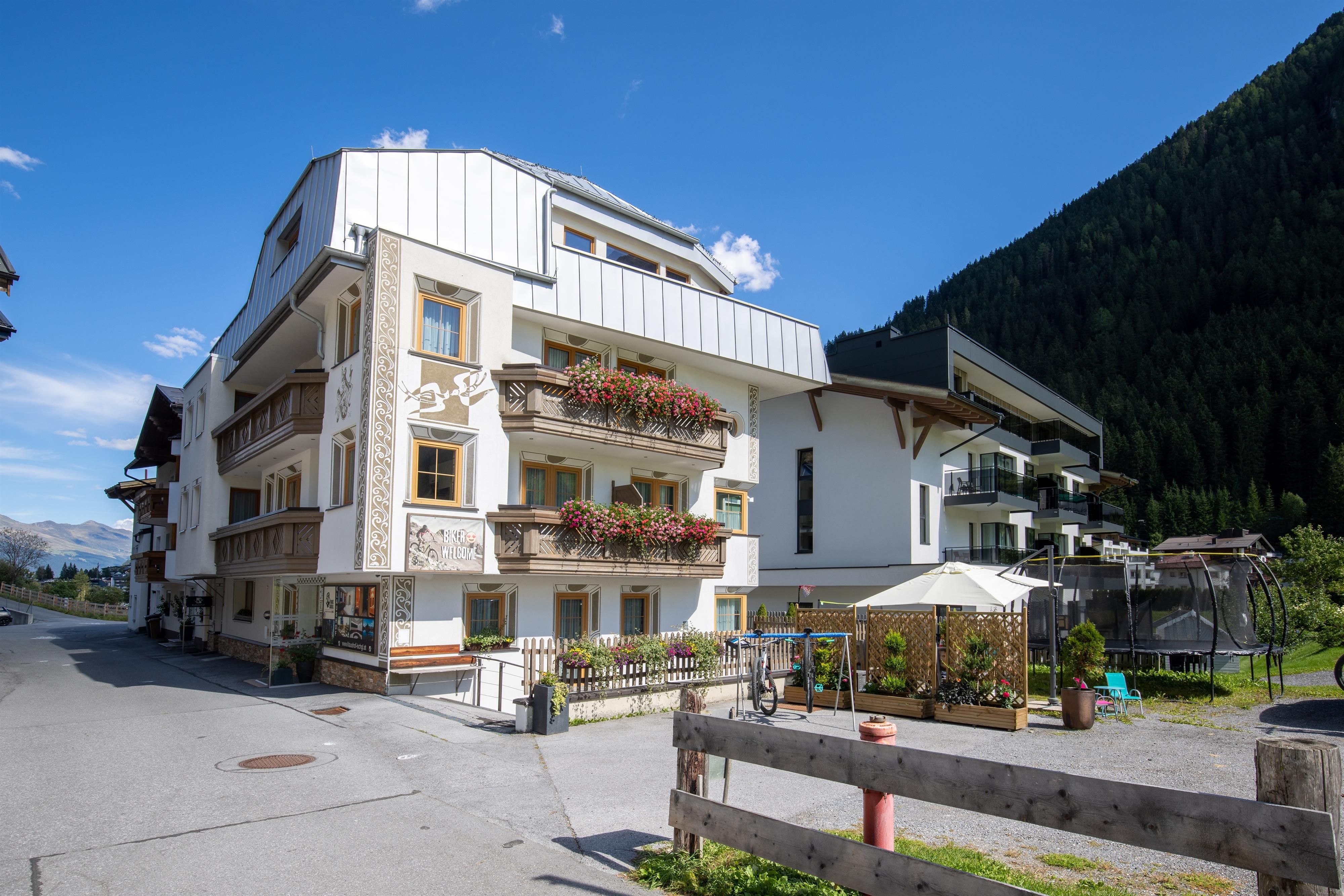 Ein modernes Gebäude mit Balkonen und Blumenarrangements. Im Hintergrund sind grüne Berge und ein blauer Himmel zu sehen.