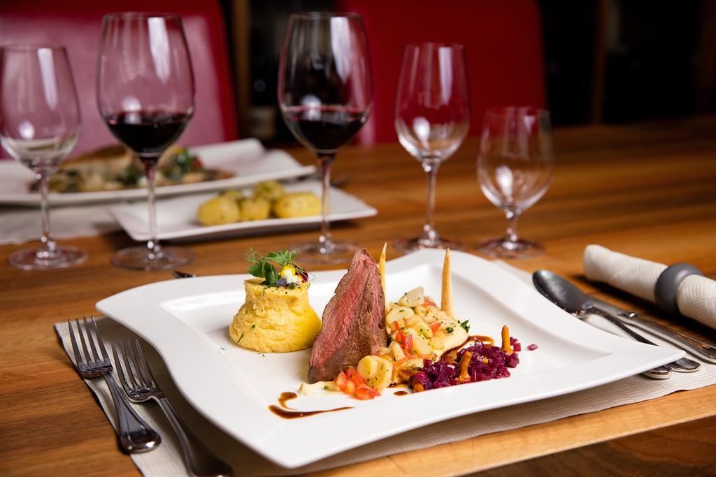 Ein elegantes Gericht auf einem weißen Teller mit Fleisch, einer Beilage aus Kartoffeln und buntem Gemüse. Im Hintergrund stehen Weingläser auf einem schön gedeckten Tisch.