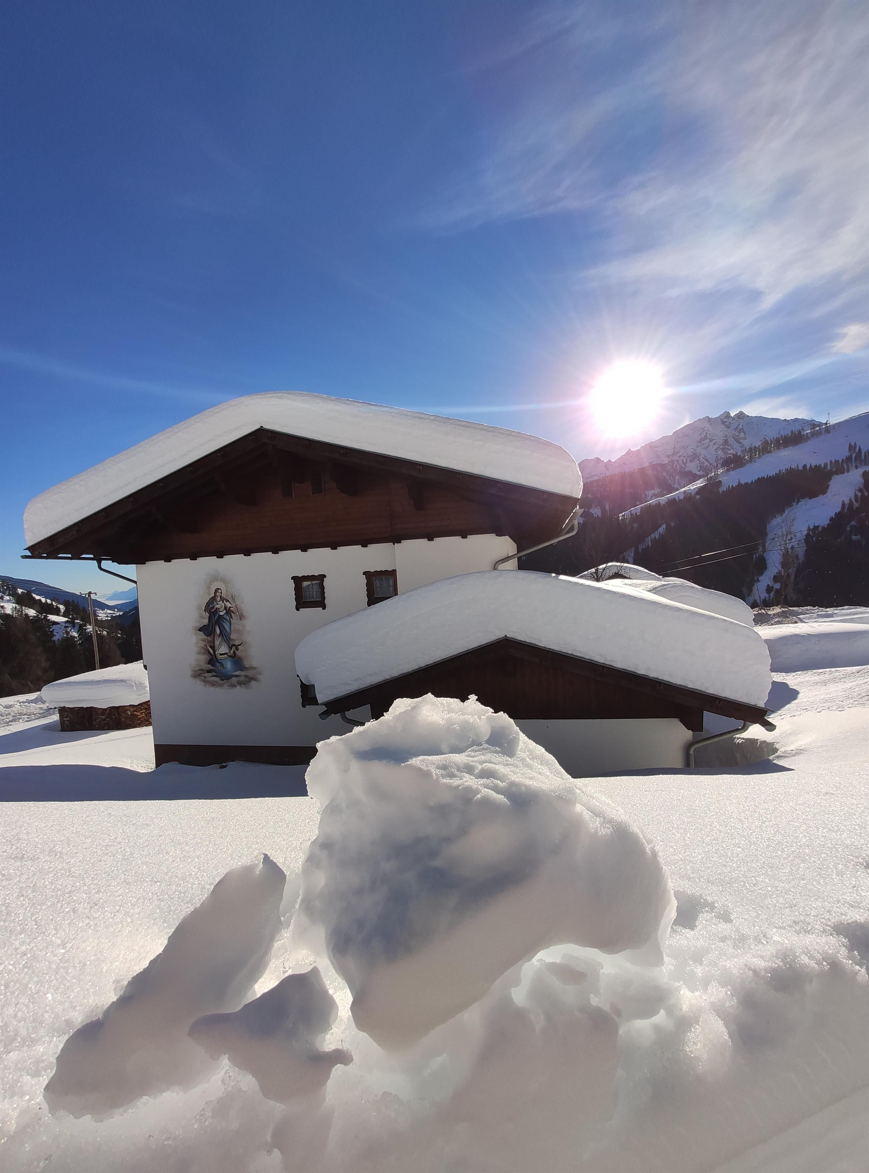 Ein charmantes Haus im Schnee mit schneebedecktem Dach und einer warmen Wintersonne. Die Umgebung ist malerisch und winterlich.