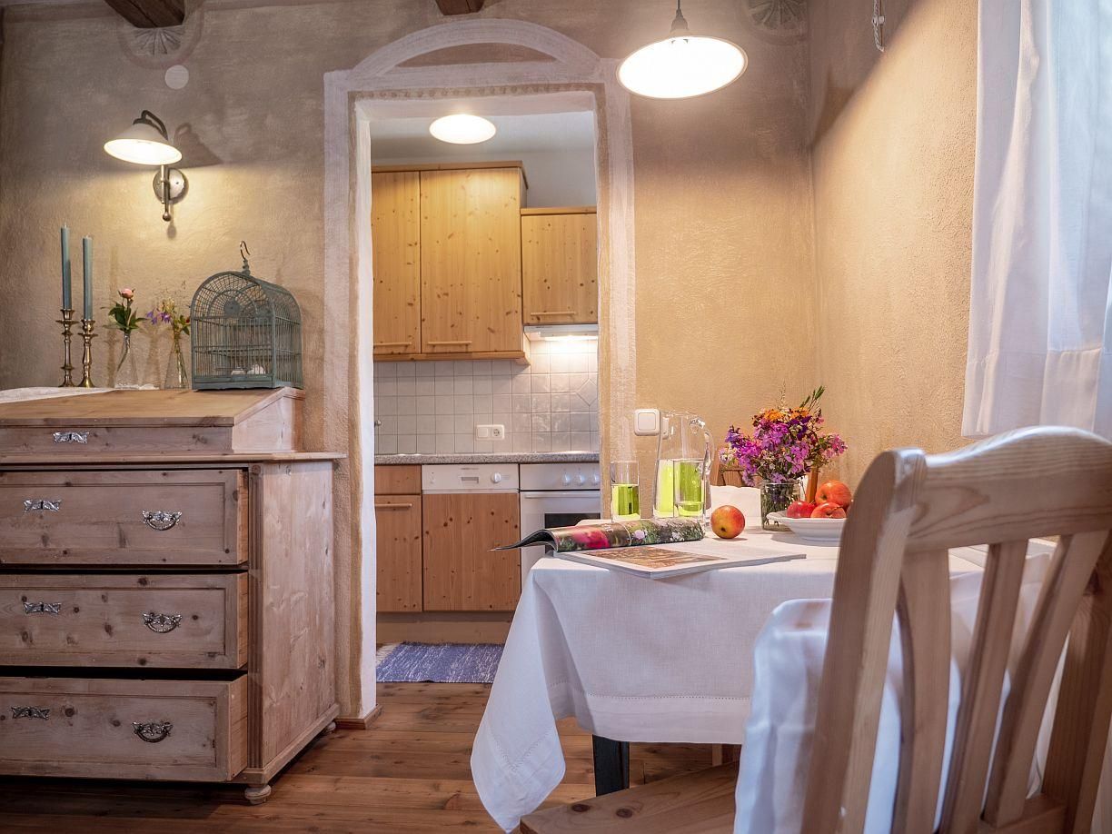 Appartement "Brotkammer" für zwei bis drei Personen geeignet, mit Holzfußboden und Holztäfelung, Durchsicht zur anschließenden Küche, weiße Vorhänge und Tischdecke, originale Holzkommode mit Deko