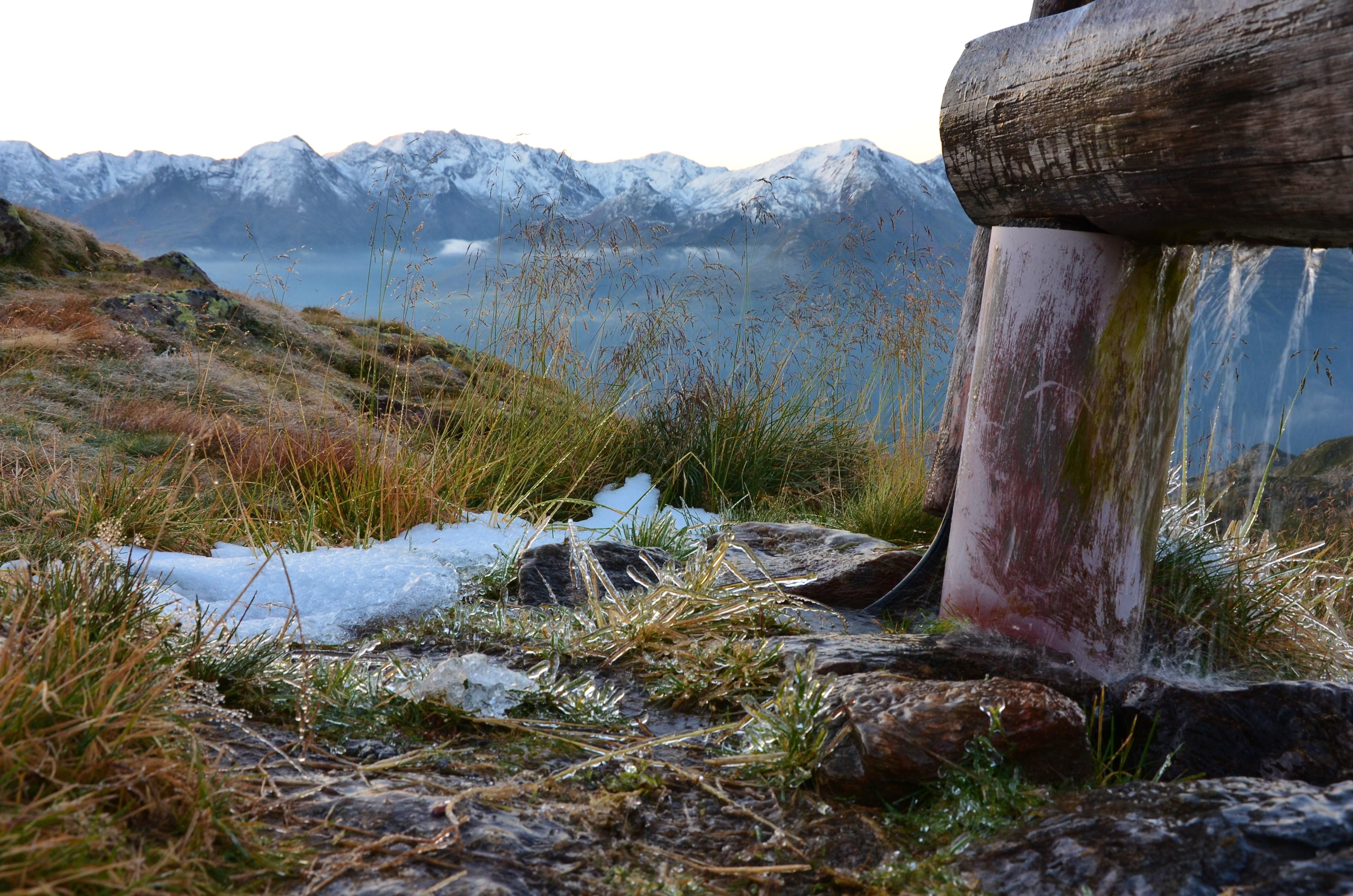 Eine alpine Landschaft mit schneebedeckten Bergen im Hintergrund. Im Vordergrund sind grüne Gräser und ein Holzpfosten zu sehen.