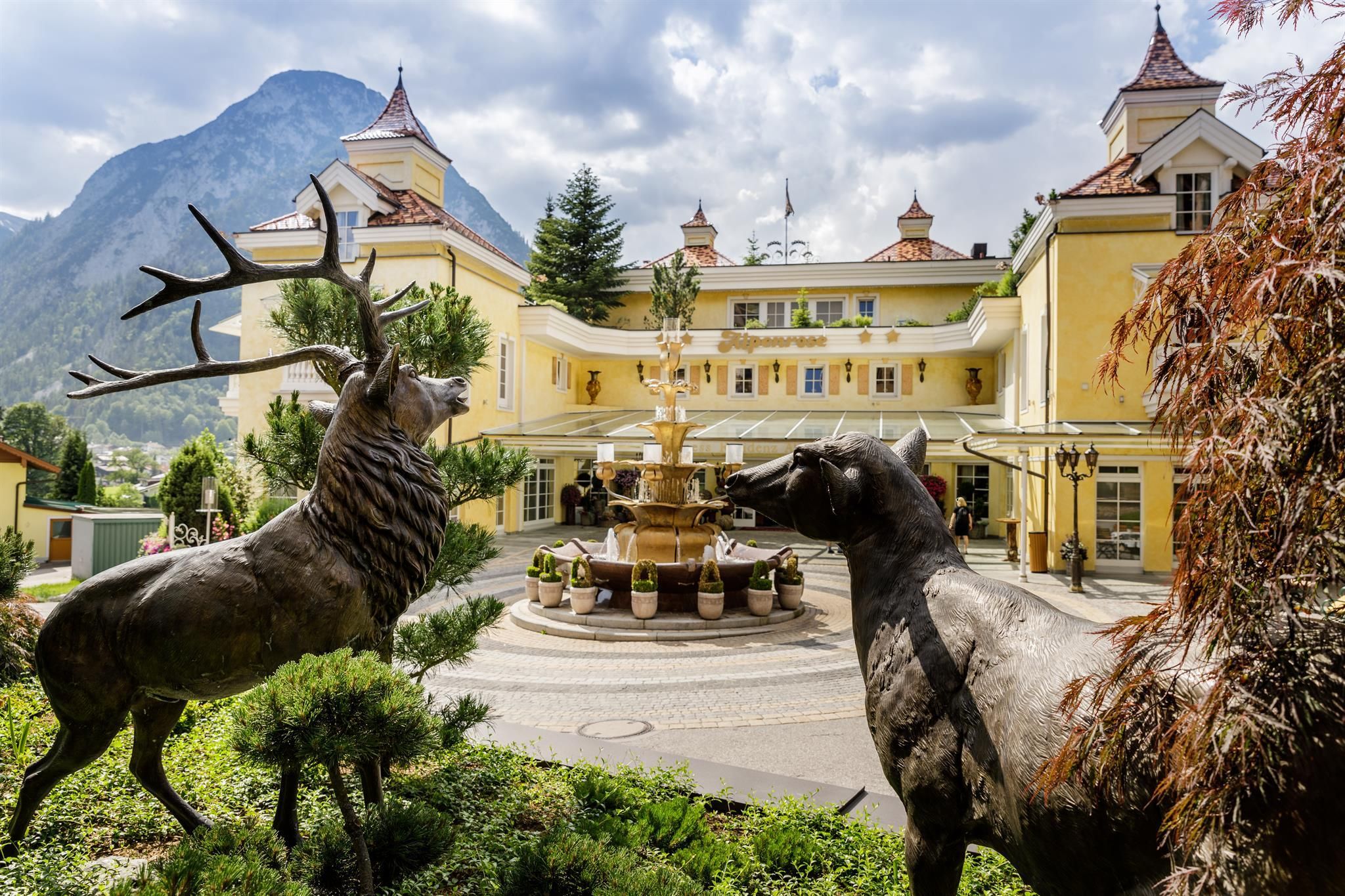 Ein malerisches Hotel mit einem Brunnen im Vordergrund. Im Garten stehen stilvolle Tierstatuen und im Hintergrund sind Berge sichtbar.