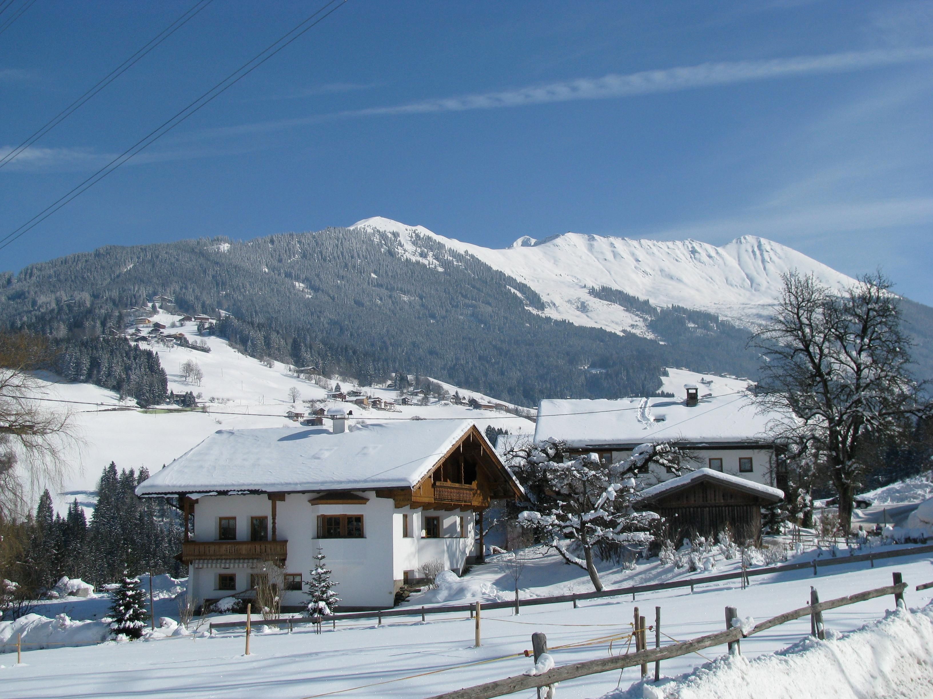 Eine verschneite Landschaft mit gemütlichen Häusern und majestätischen Bergen im Hintergrund. Der klare Himmel und die strahlende Sonne verleihen der Szene eine friedliche Atmosphäre.