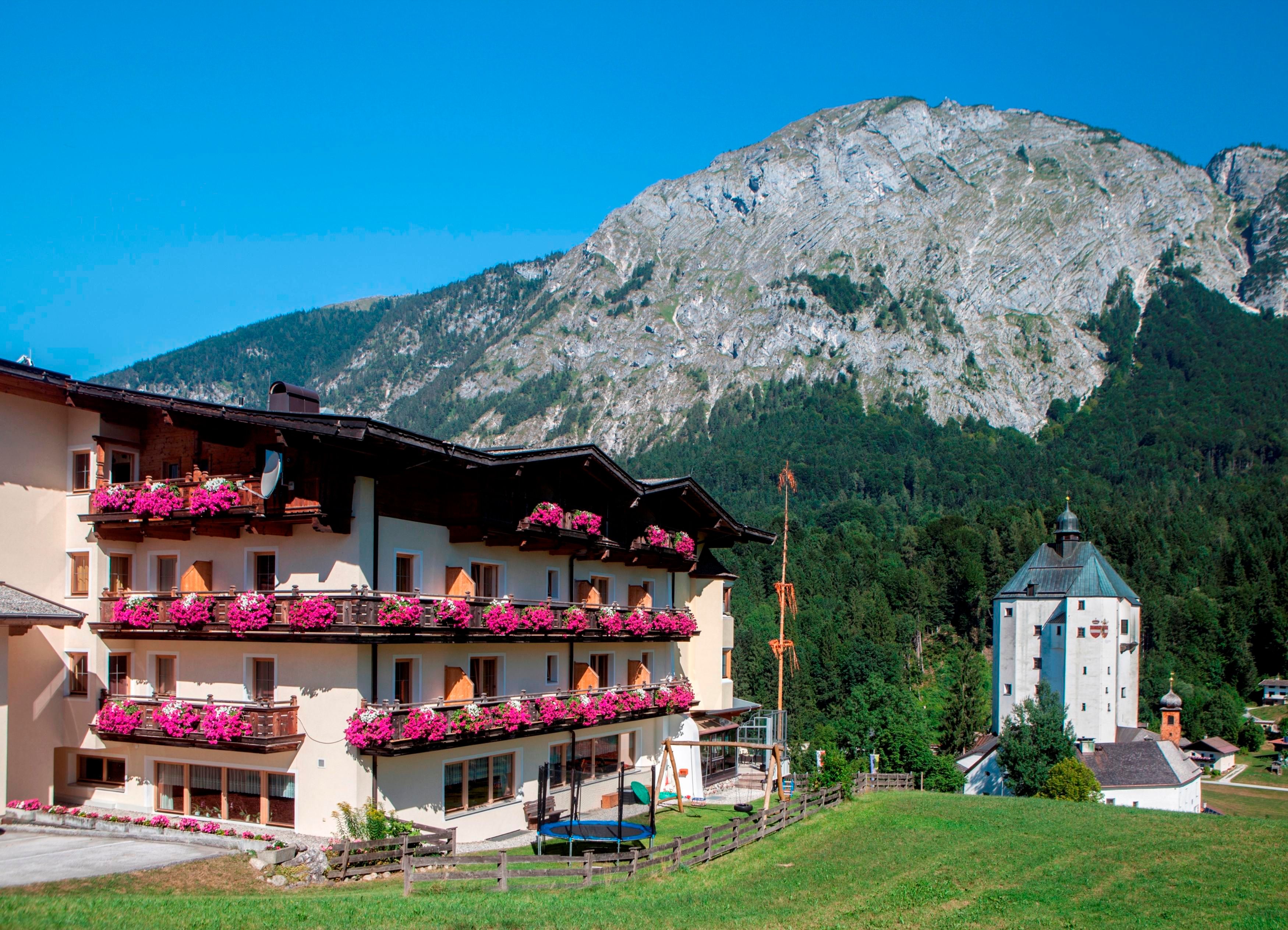 Ein malerisches Hotelgebäude mit Blumenbalkonen steht vor einer beeindruckenden Bergkulisse. Der klare Himmel und die grüne Umgebung schaffen eine einladende Atmosphäre.