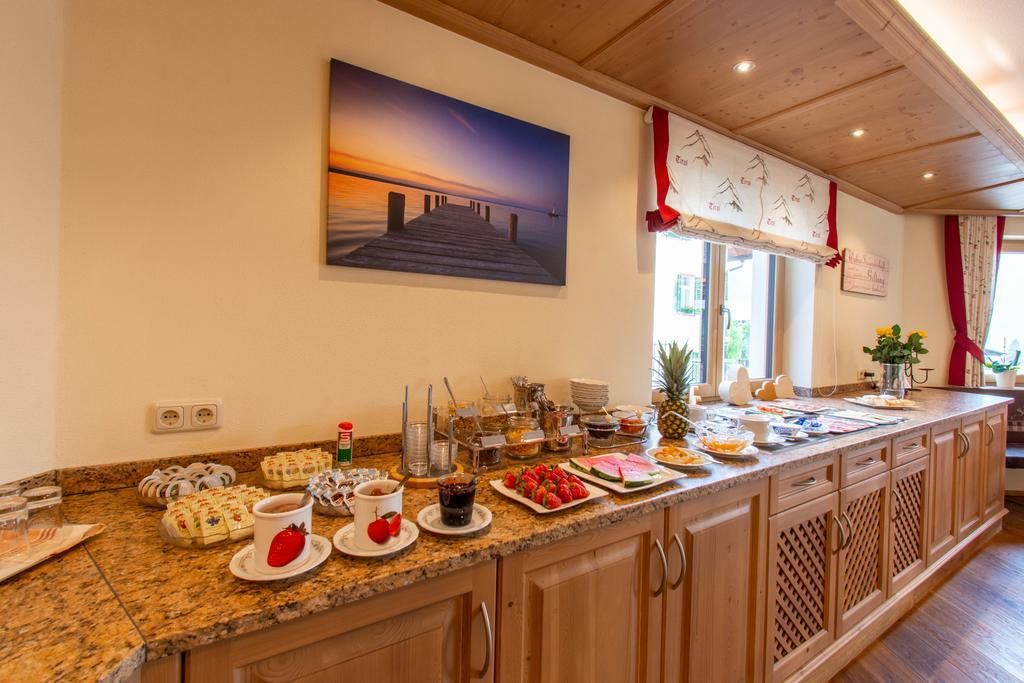 Eine einladende Frühstückslandschaft mit einer Vielzahl von Speisen und Getränken auf einer Holztheke. Im Hintergrund hängt ein Bild eines Sonnenuntergangs über einem Steg.