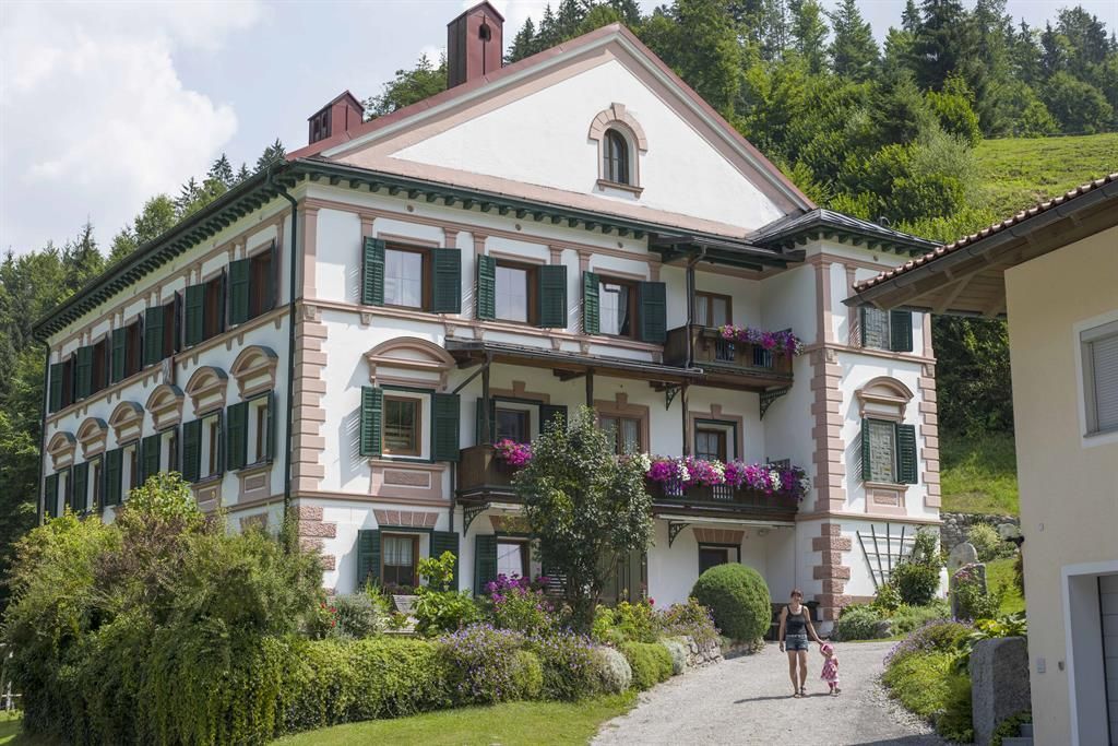 Ein großes, elegantes Gebäude mit bunten Blumenbalkonen und grünen Fensterläden. Eine Person geht auf einem gepflasterten Weg in Richtung des Hauses.