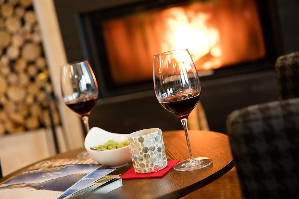 Zwei Gläser Rotwein stehen auf einem Tisch neben einer Schüssel mit Snacks. Im Hintergrund brennt ein gemütlicher Kamin.