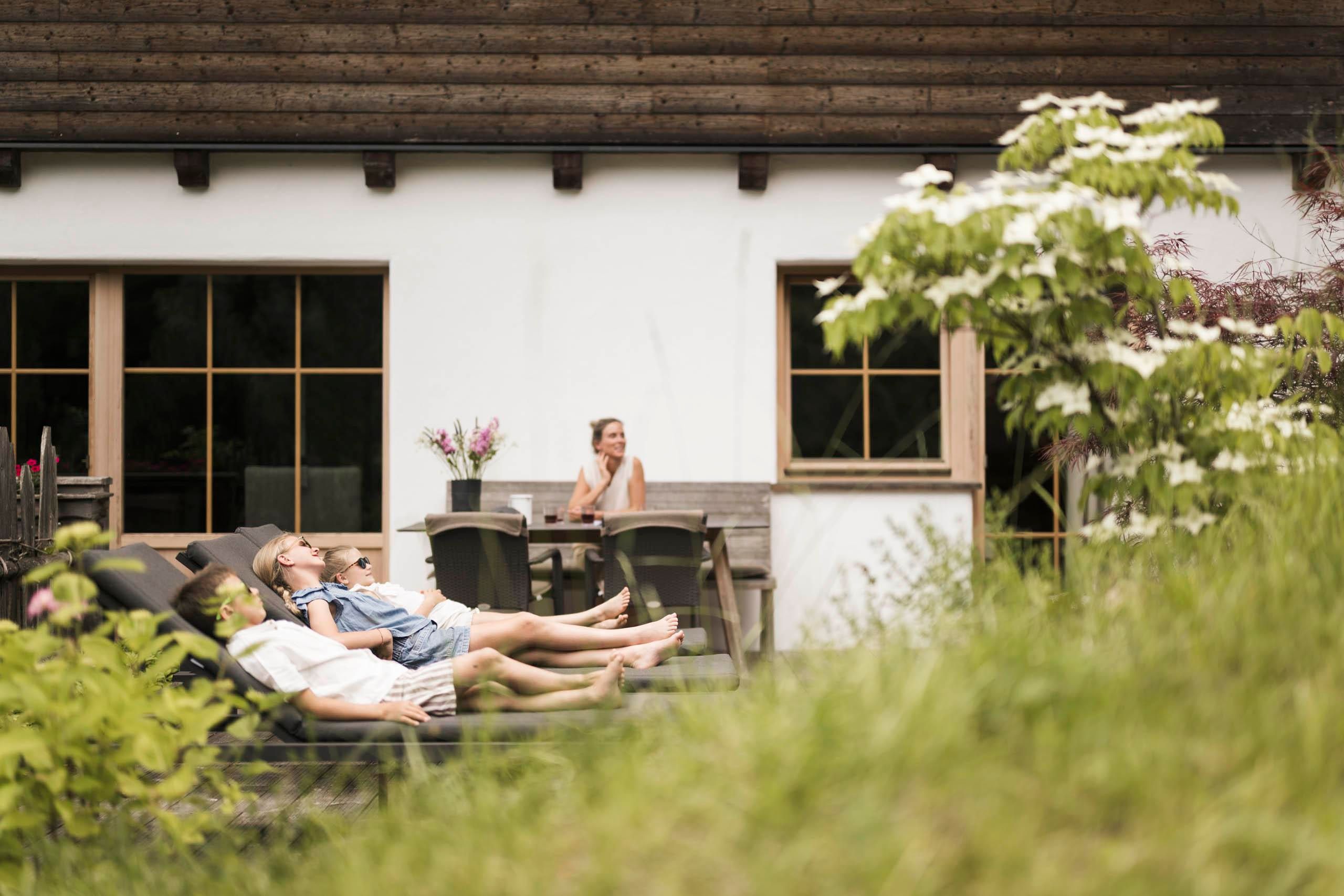Eine Familie entspannt auf Liegen im Garten eines Hauses. Im Hintergrund ist eine Frau zu sehen, die in einem Stuhl sitzt und die Umgebung genießt.