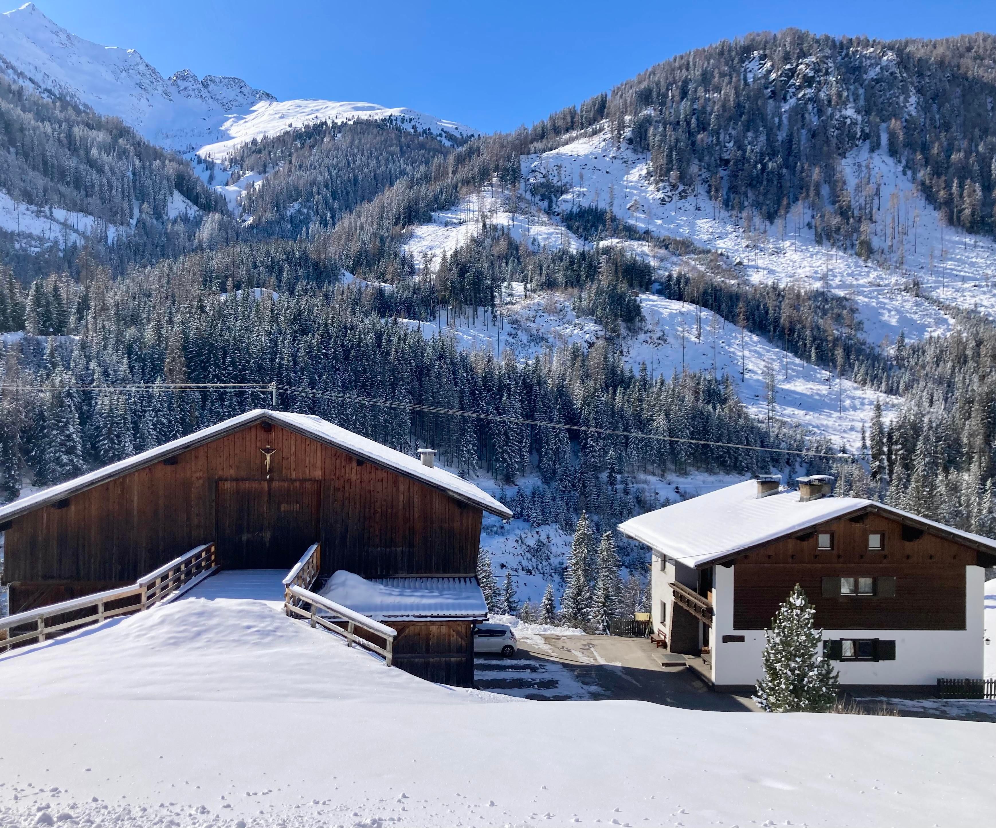 Hof und Haus inmitten einer idyllischen Winterlandschaft. Dahinter ragen Berge vor einem klaren, blauen Himmel auf.